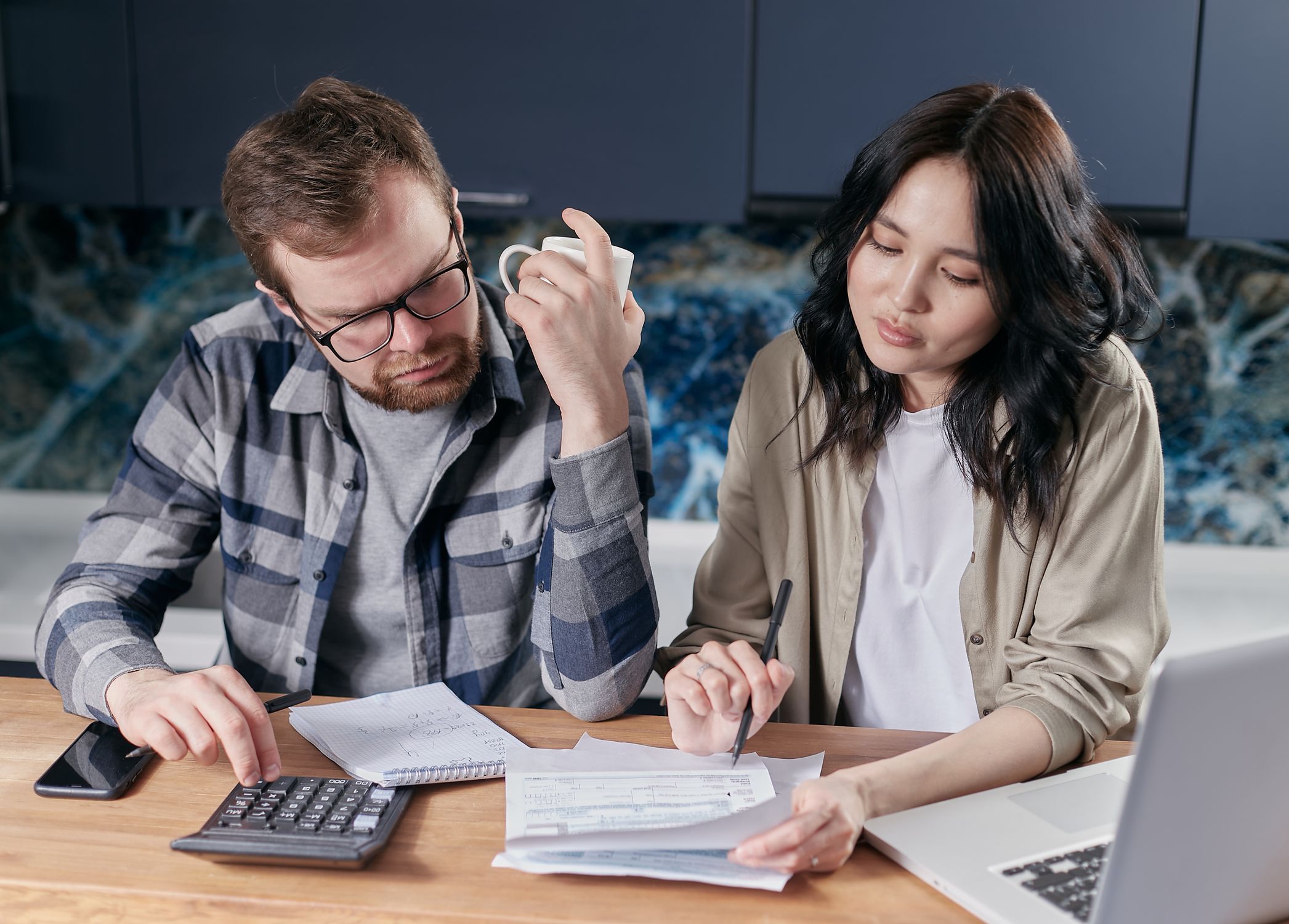 A couple reviewing household bills with a calculator and laptop at a kitchen table.