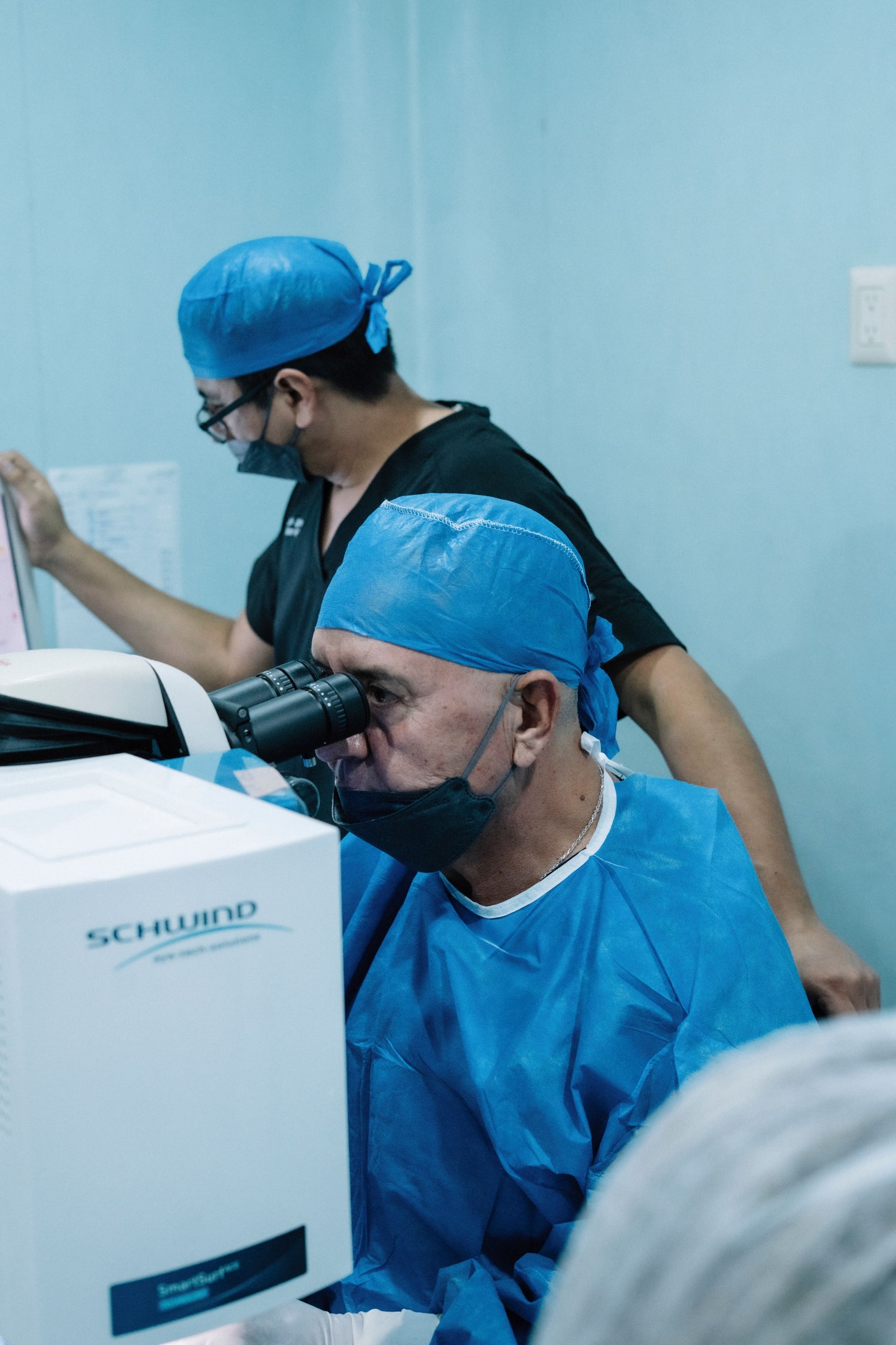 A surgeon looks through an eye-surgery microscope while another clinician stands behind him.