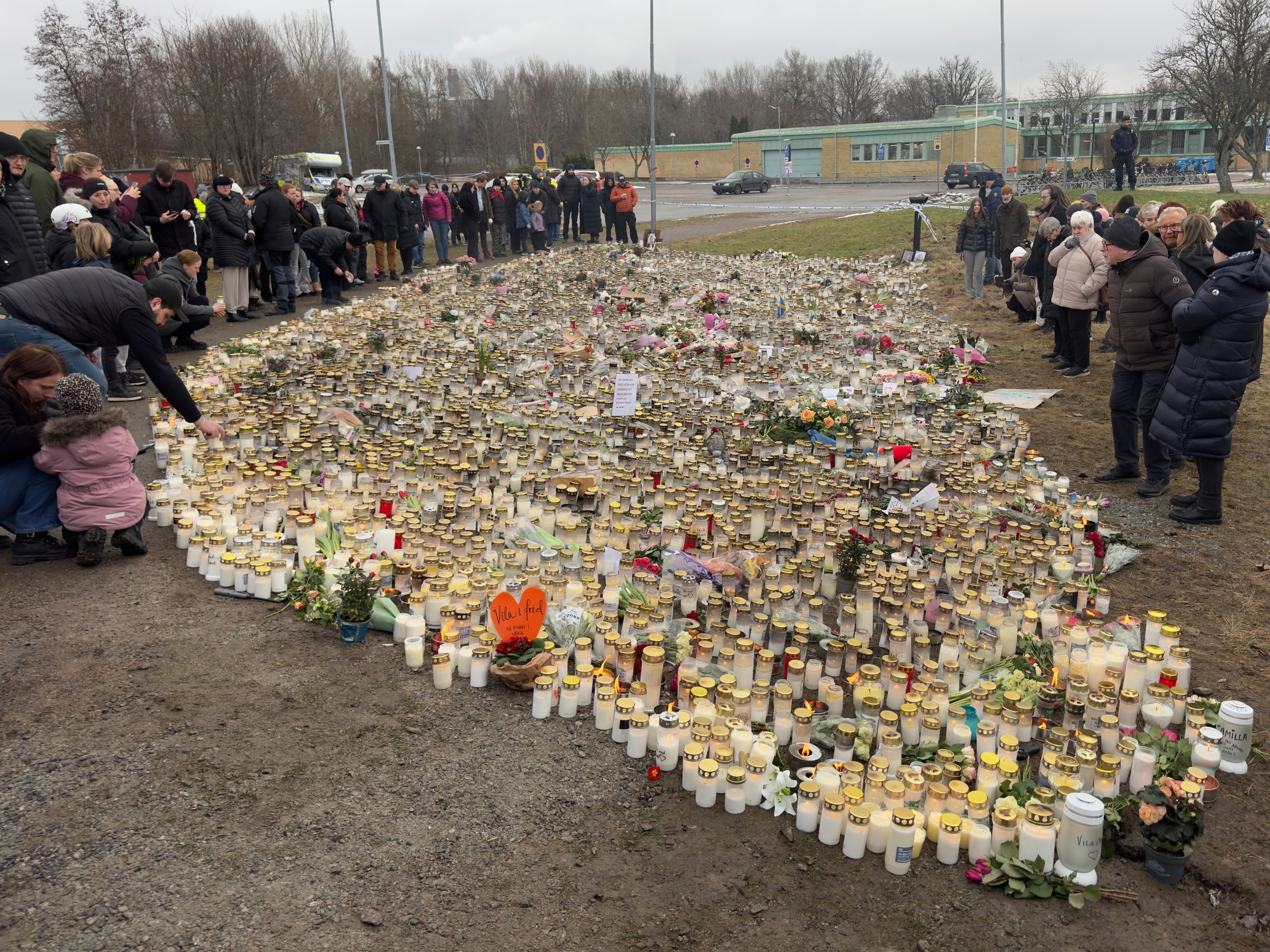 Candles and flowers cover the memorial outside Campus Risbergska as people stand around it.