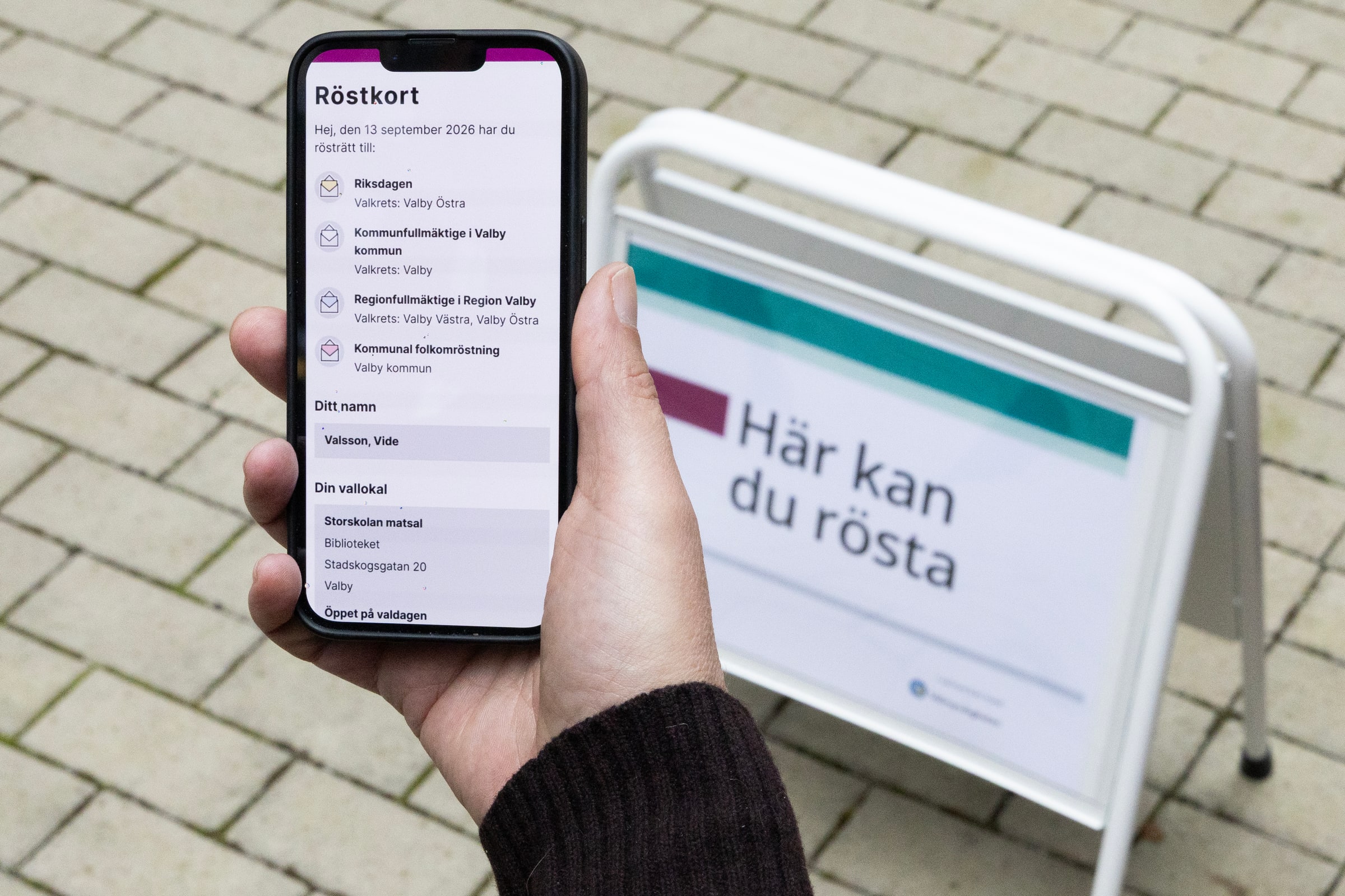 A hand holds a phone displaying a digital voting card beside a Swedish polling-place sign.
