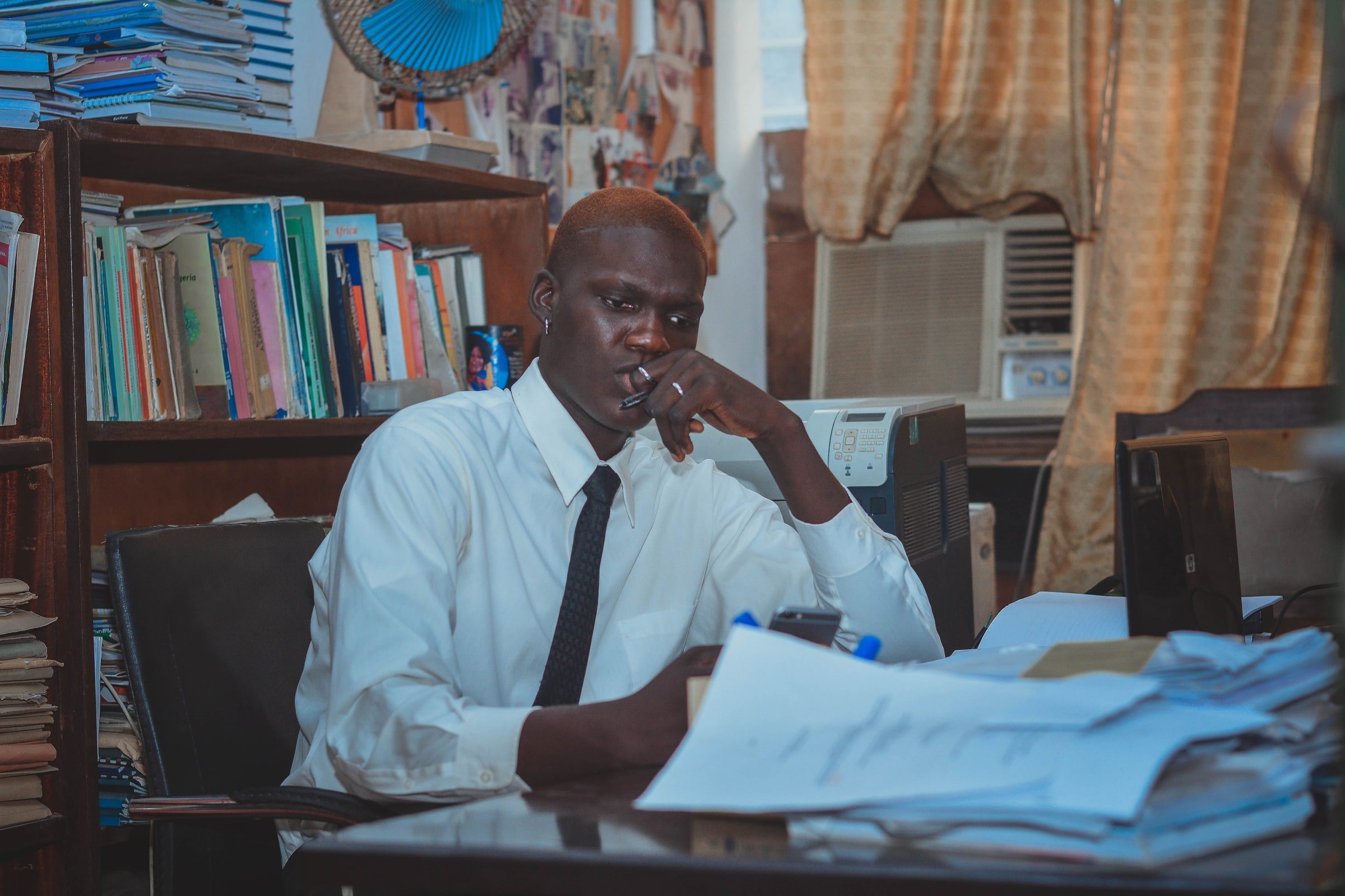 A man holds a pen near his mouth at an office desk covered in papers, beside a laptop.