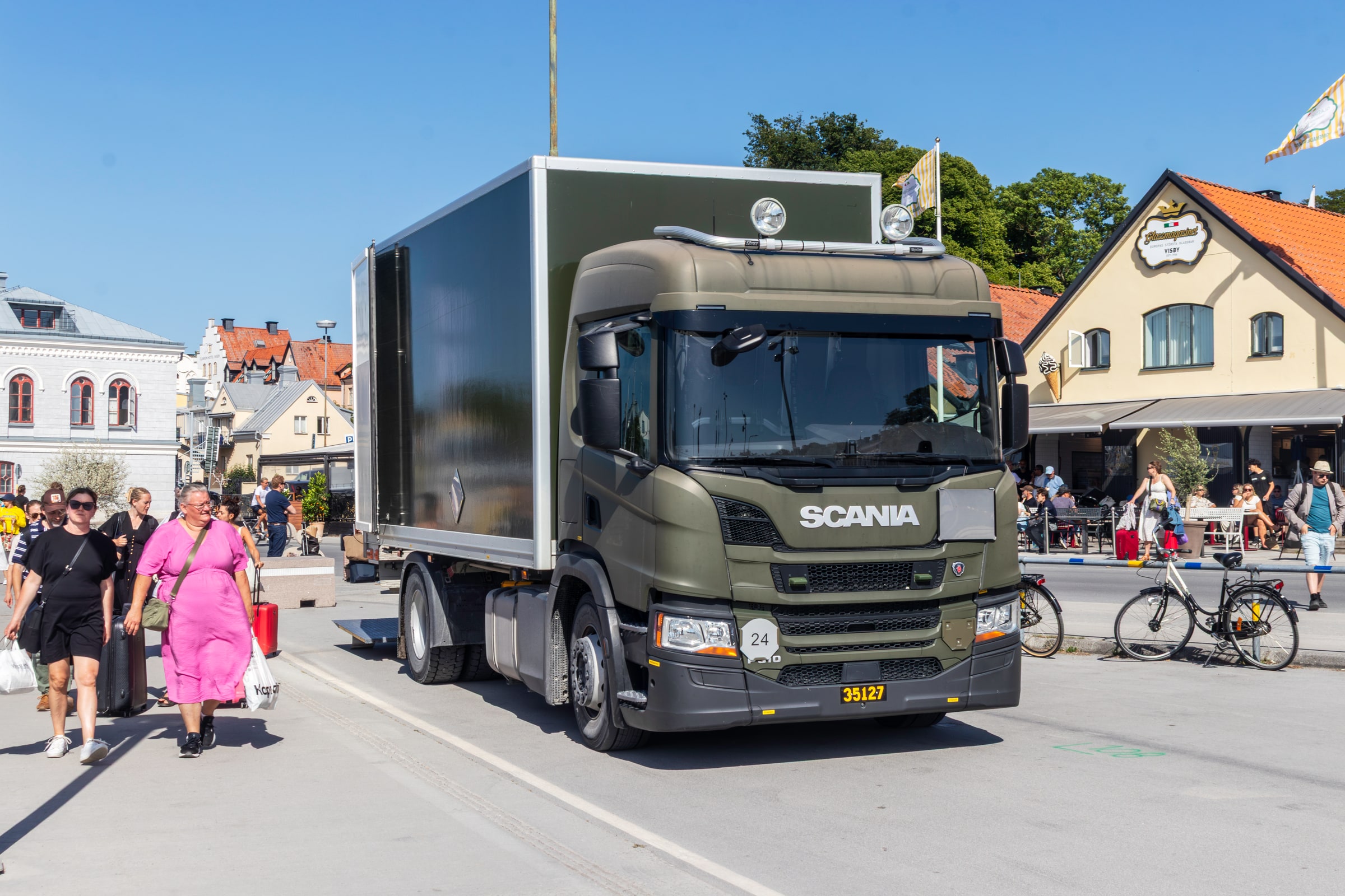A green Swedish army box truck drives along a street in Visby beside pedestrians.