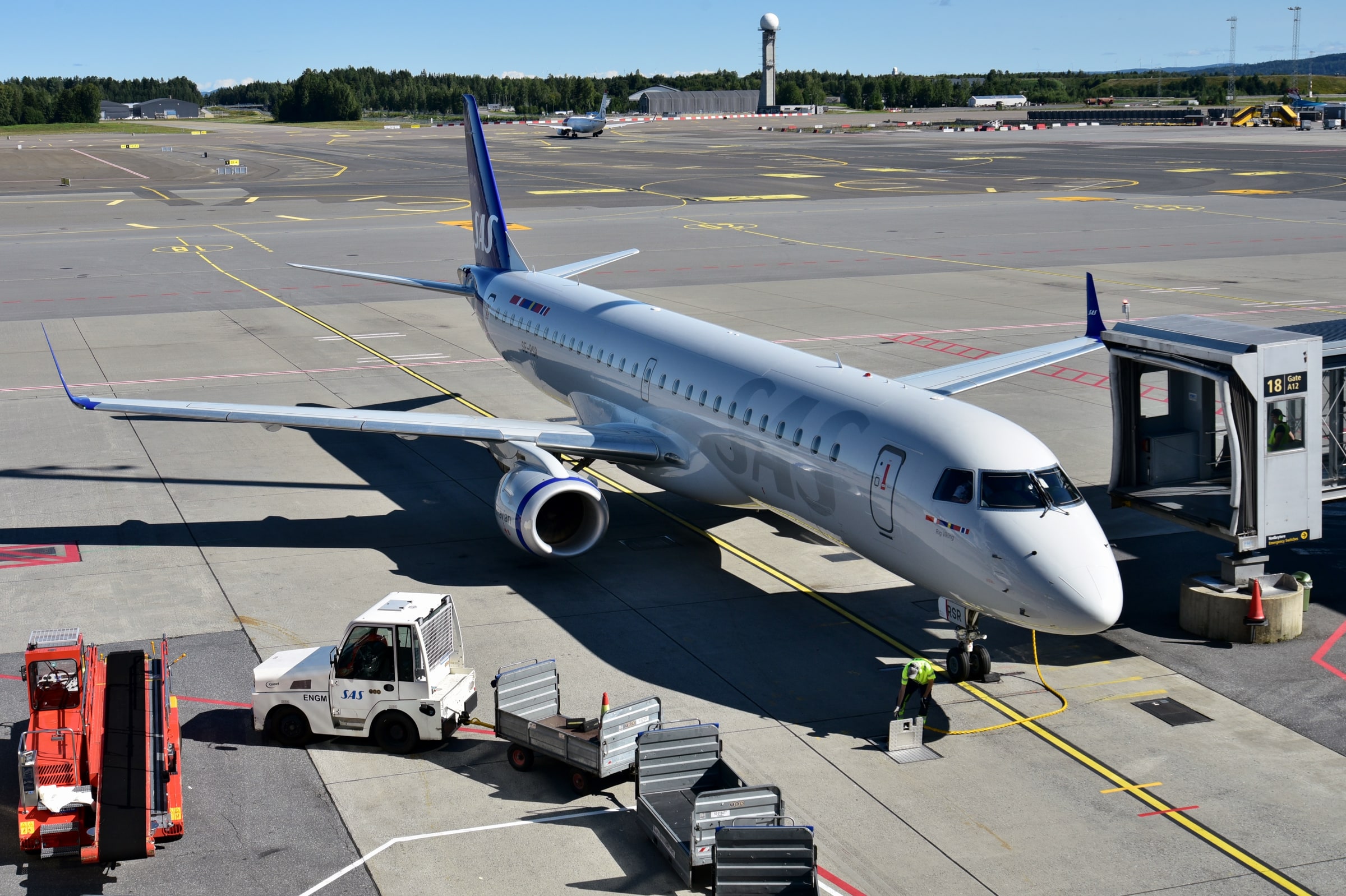 A SAS Link Embraer 195 beside a jet bridge and baggage equipment at Oslo Airport in August 2024.