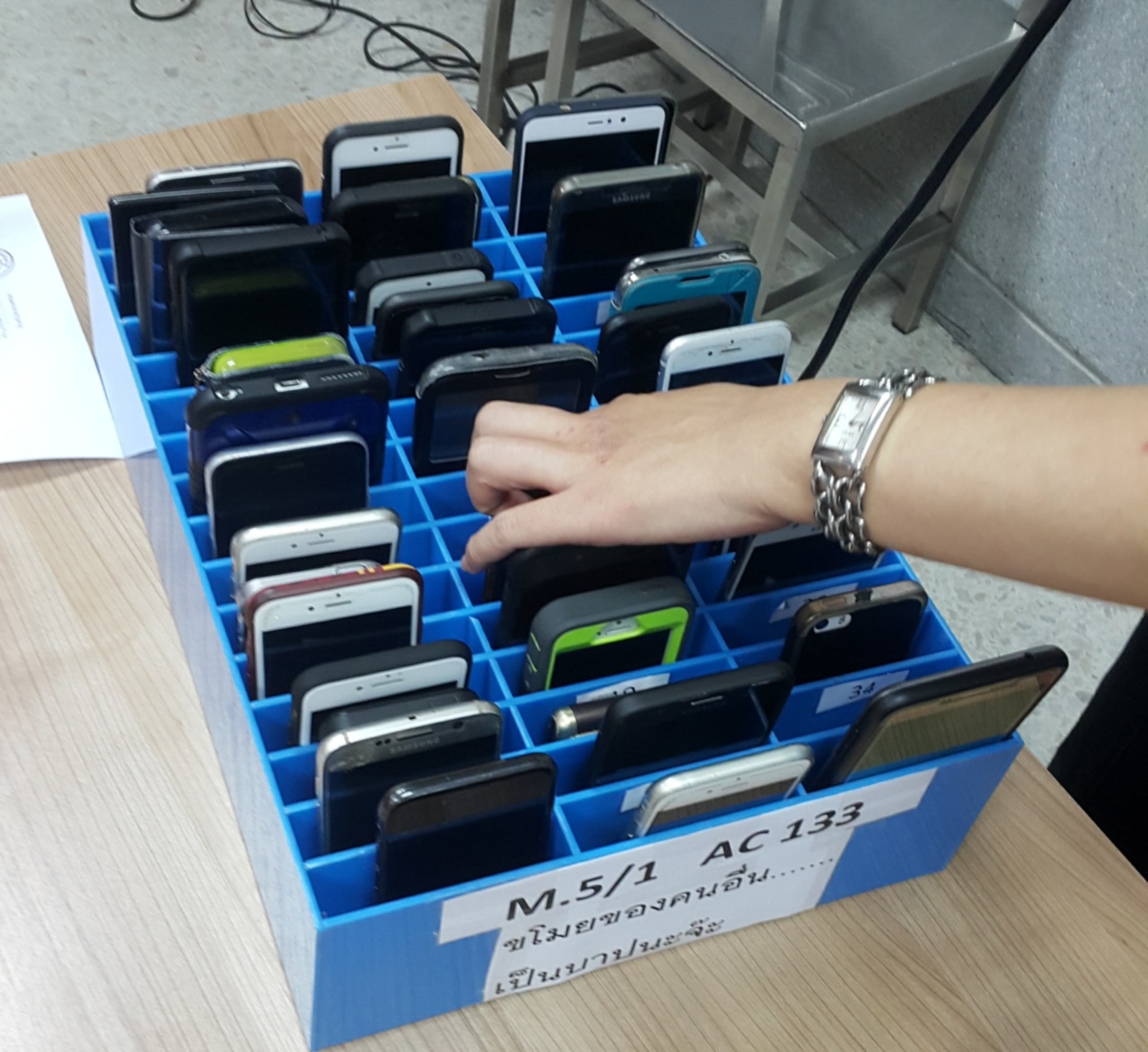 A hand reaches into a blue classroom storage cage filled with students' mobile phones.
