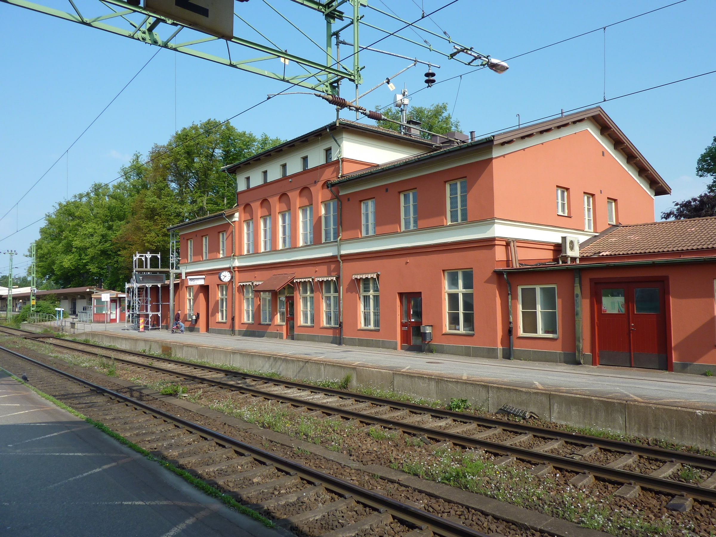 Alingsås station's red building beside railway tracks and overhead power lines on a clear summer day.