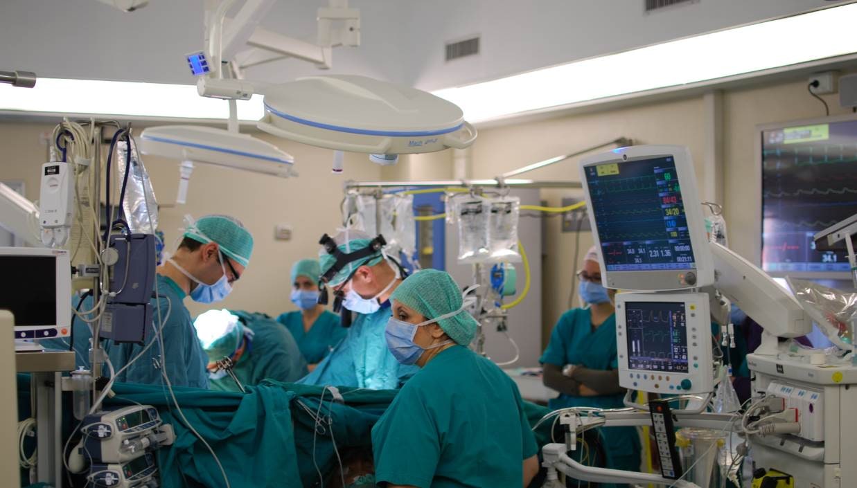 A masked cardiac surgical team works around a covered patient area with monitors and clinical equipment.