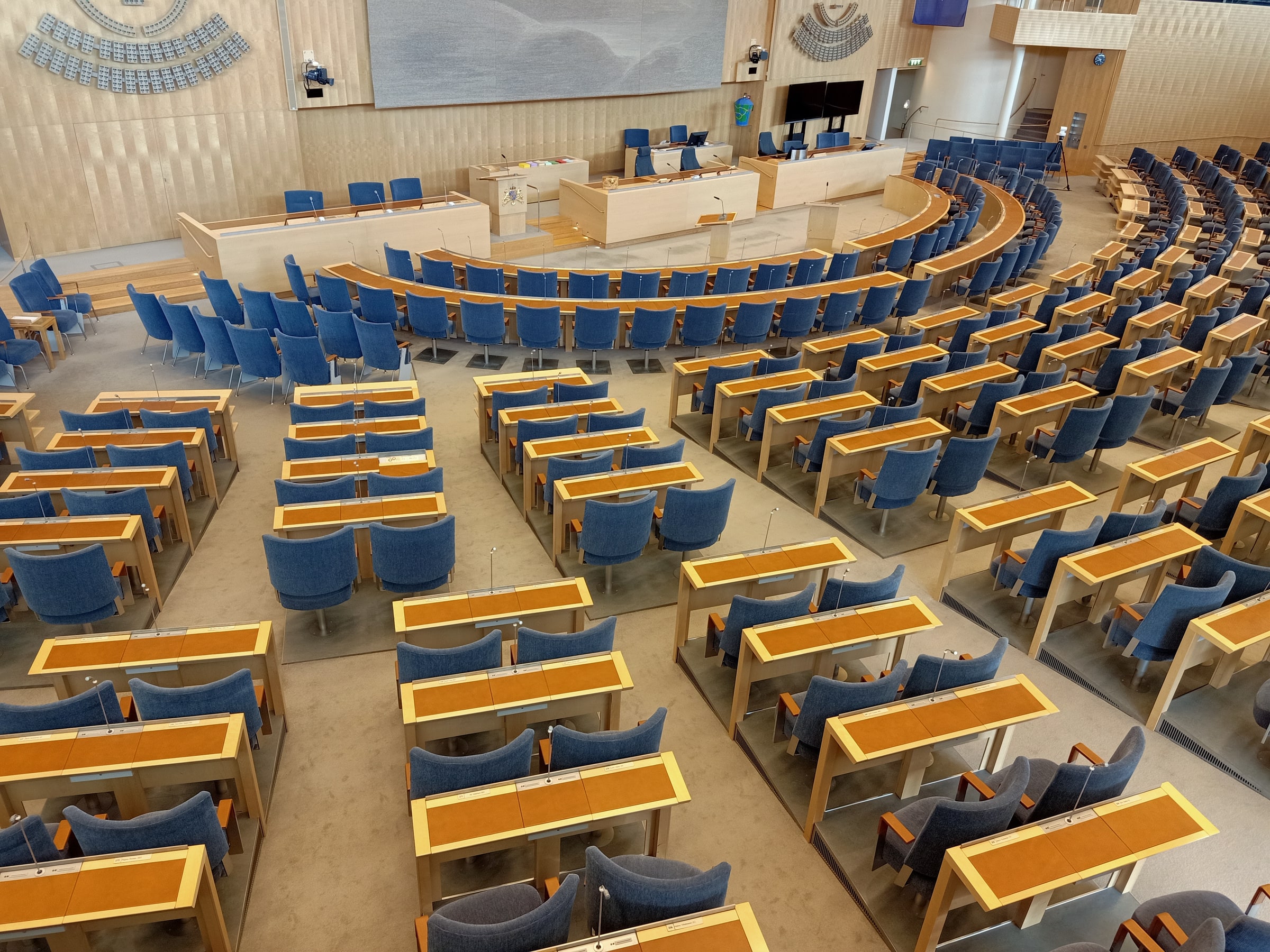 Rows of individual desks and blue seats face the front benches of the Swedish Parliament chamber.