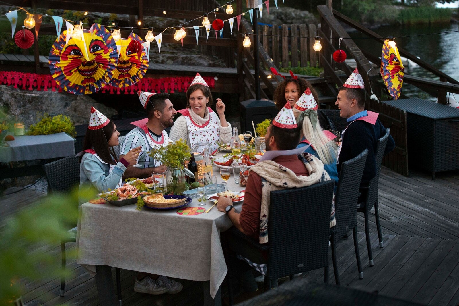 Seven people wearing paper hats and bibs share crayfish at a decorated waterside table.