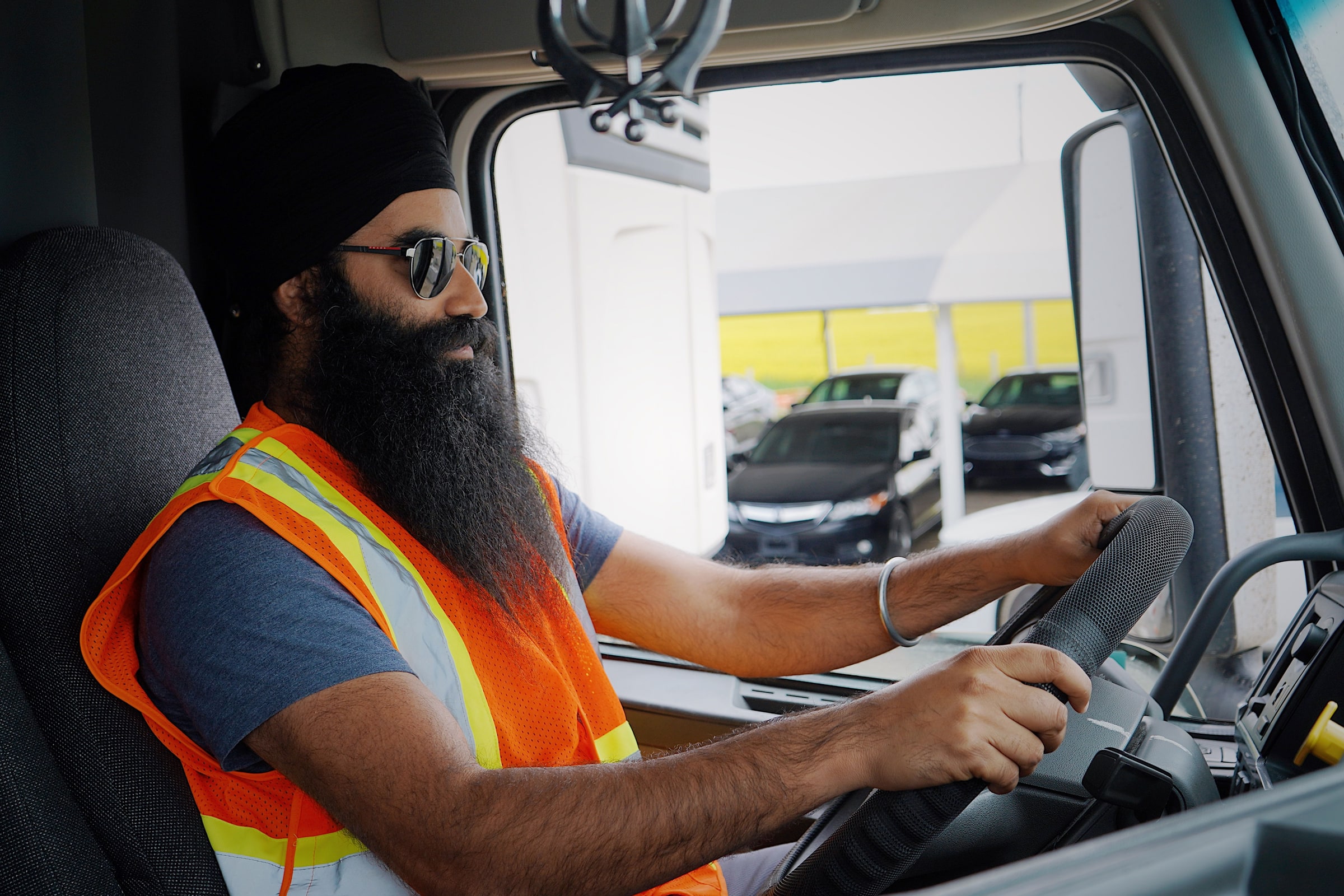 A truck driver in a safety vest works at the wheel inside a lorry cab.