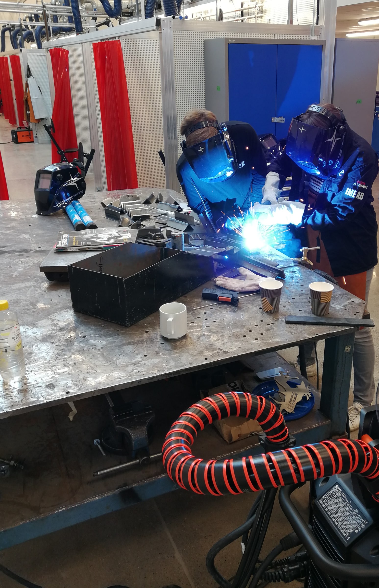 Student and teacher wearing welding masks at a metal workbench. Bright welding arc, gloved hands, metal pieces and extraction hose show practical training.
