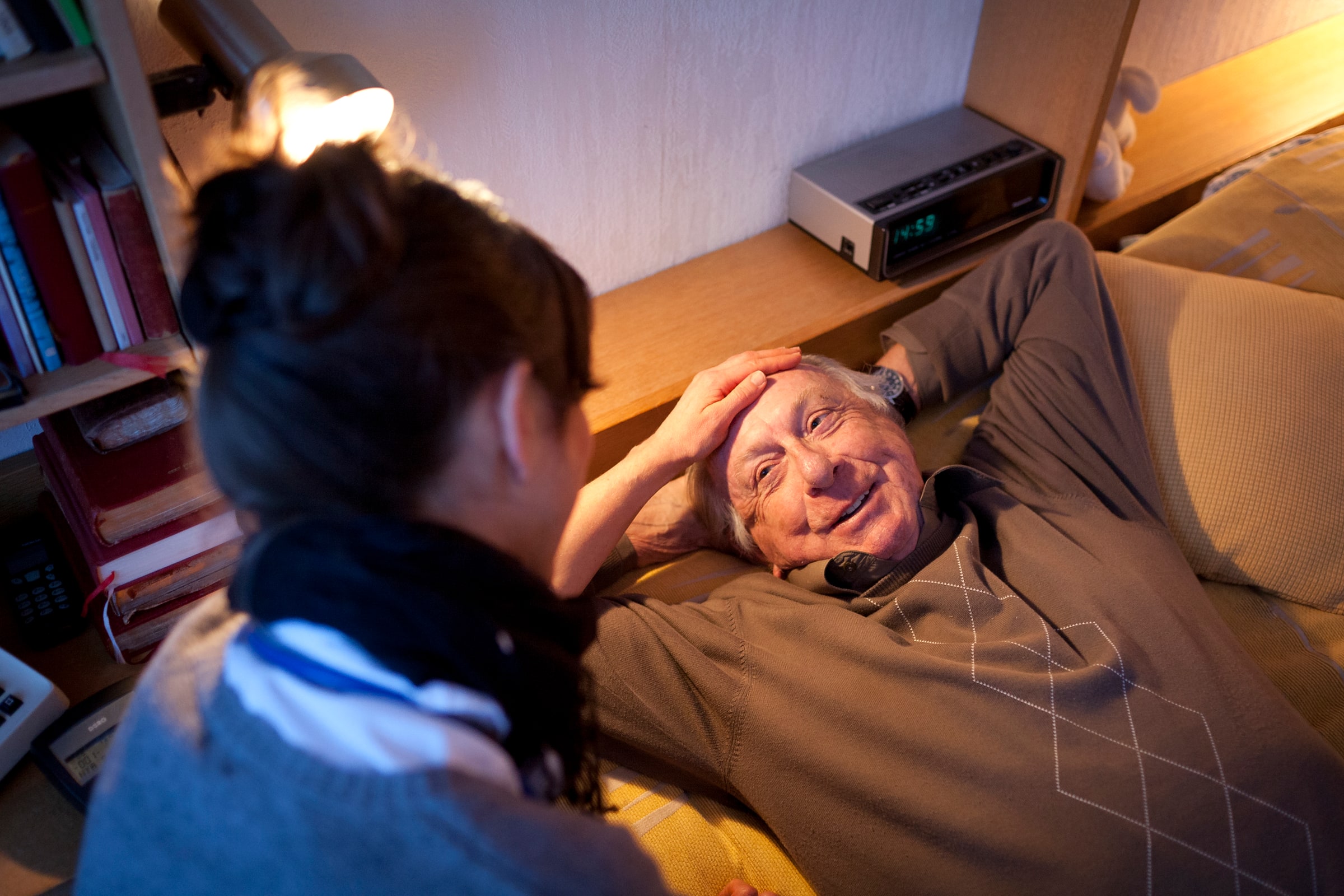 A care worker talks with an older man reclining on a sofa at home.