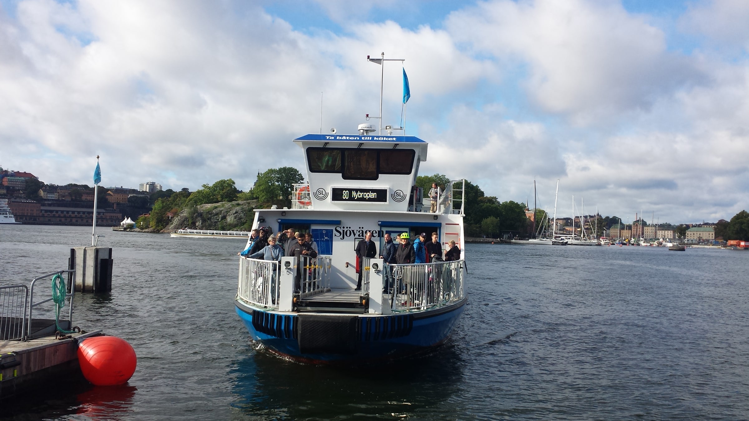 Passengers stand on the bow of the blue-and-white Sjövägen ferry approaching a landing in Stockholm.