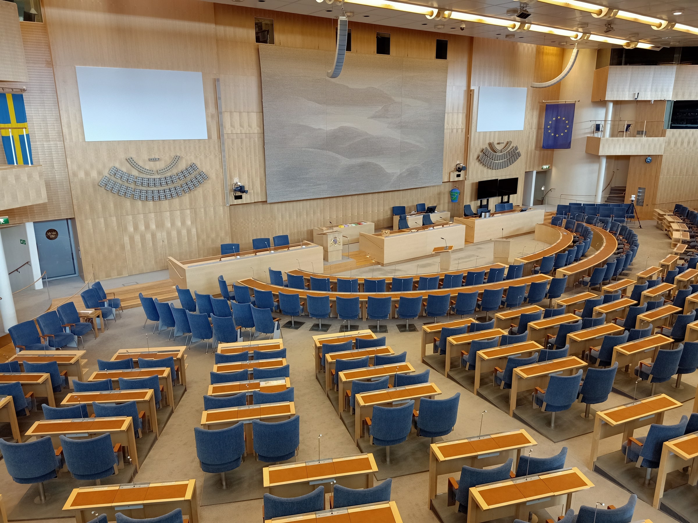 Curved blue seating and desks face the speaking platform in the Swedish Parliament chamber.