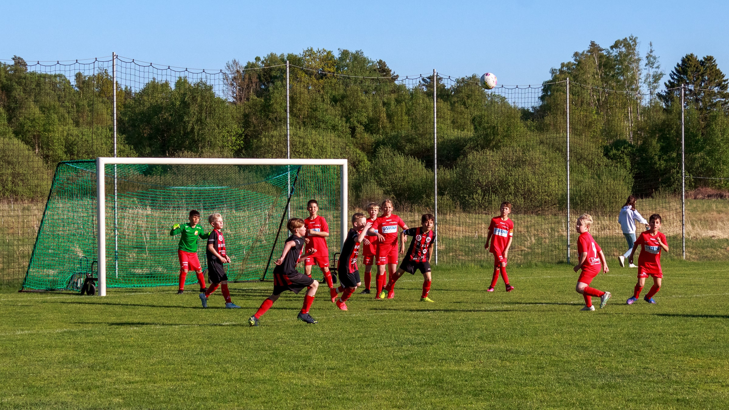 Children in red and black kits play football in front of a goal at Brastad Arena.