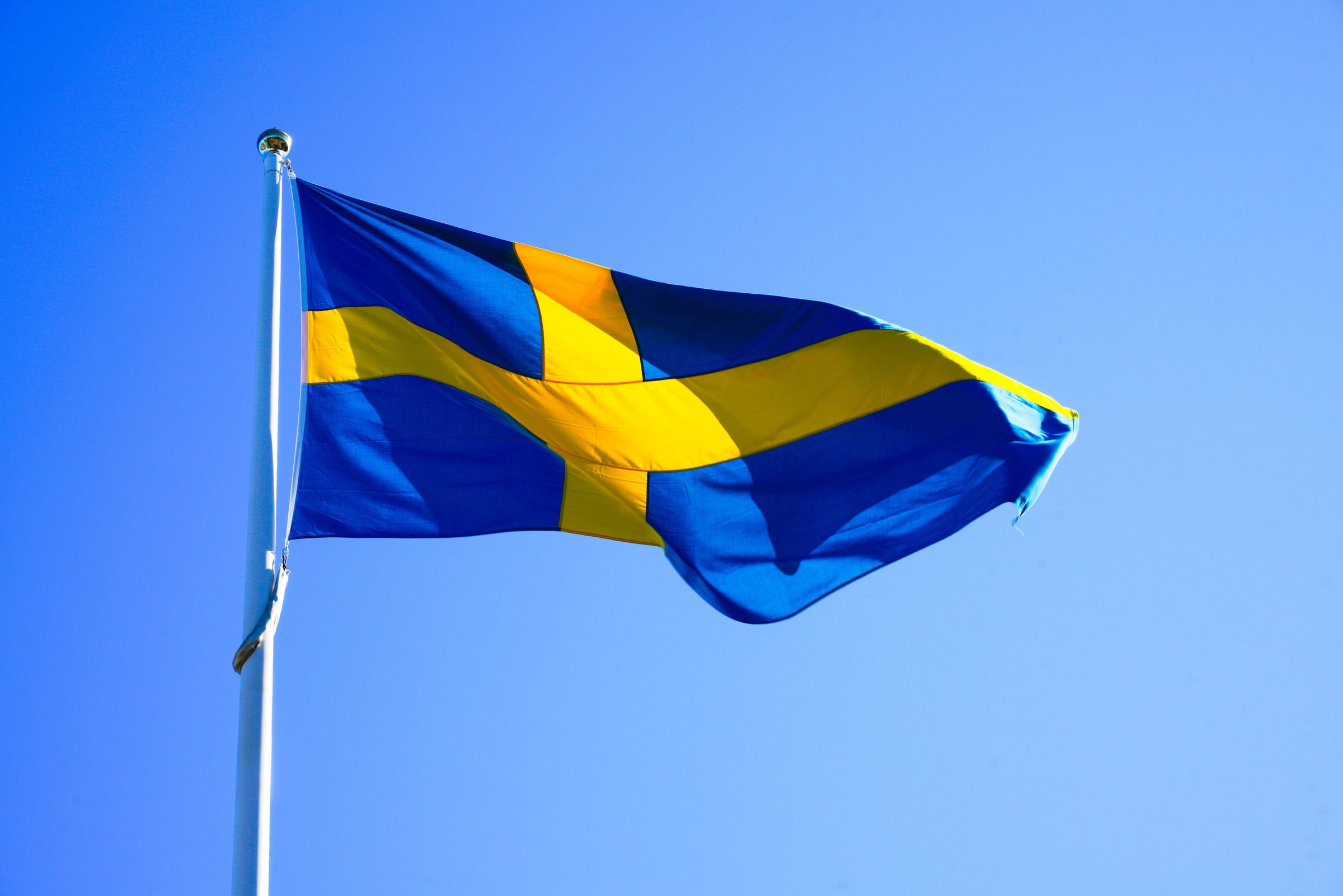 A Swedish flag waves from a white flagpole against a clear blue sky.