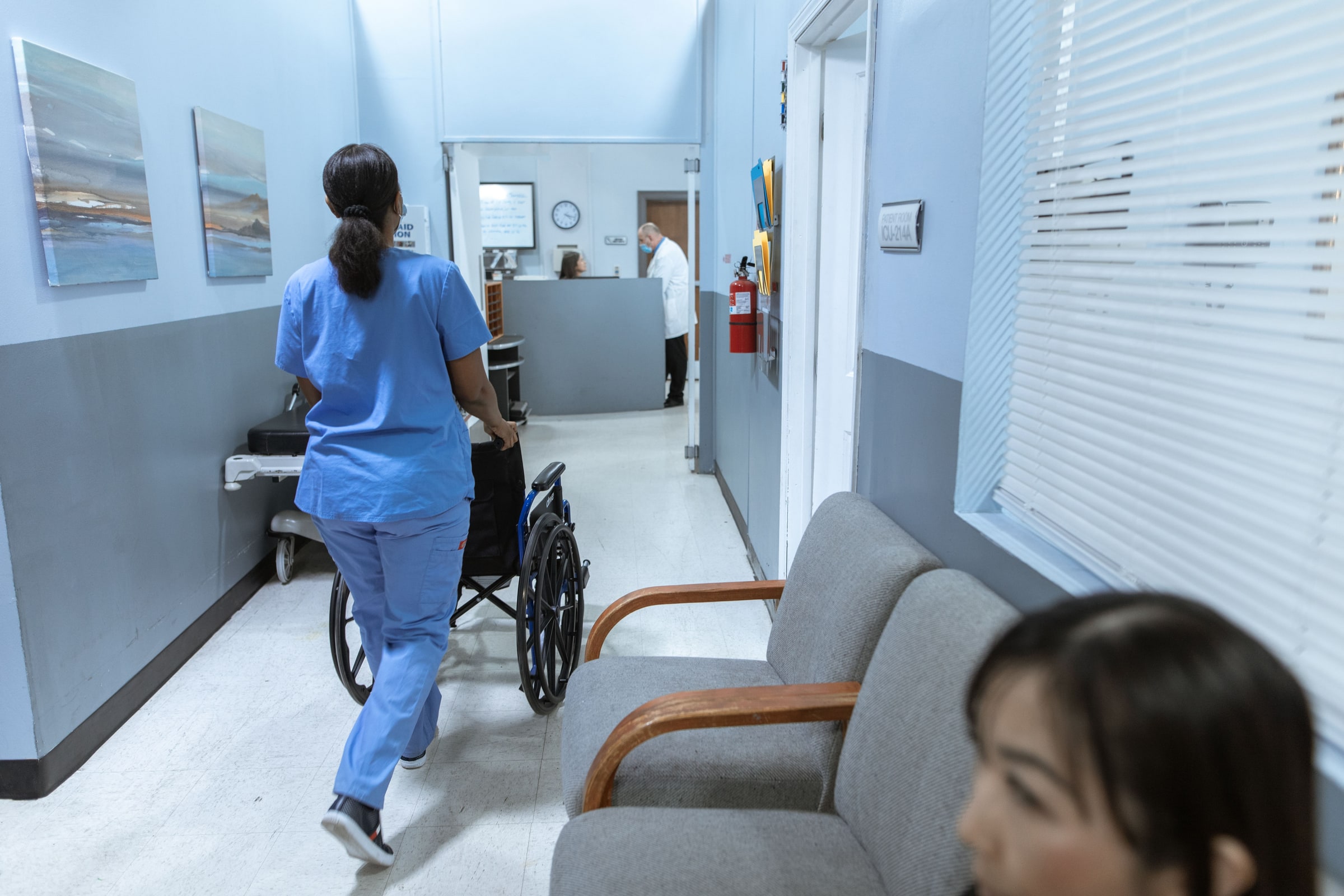 A care worker in blue scrubs pushes a person in a wheelchair through a clinical corridor.