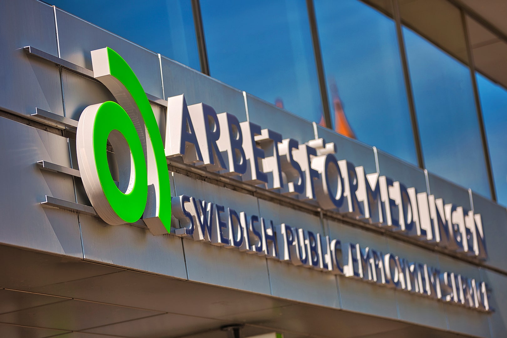 A close view of the green Arbetsförmedlingen symbol and bilingual agency name on a glass-fronted building.
