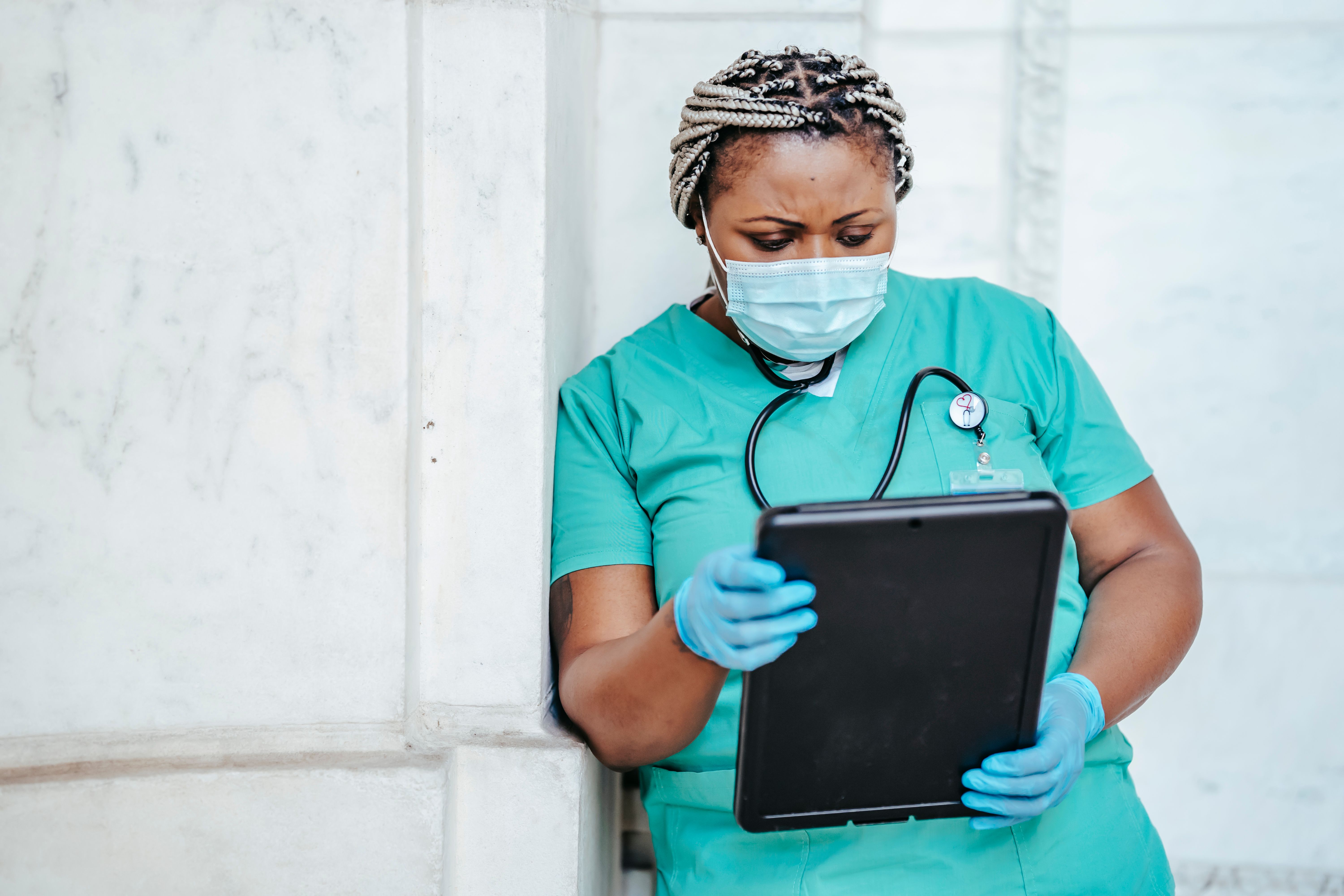 A Black woman in blue scrubs and protective equipment reviews documents while working in a hospital.