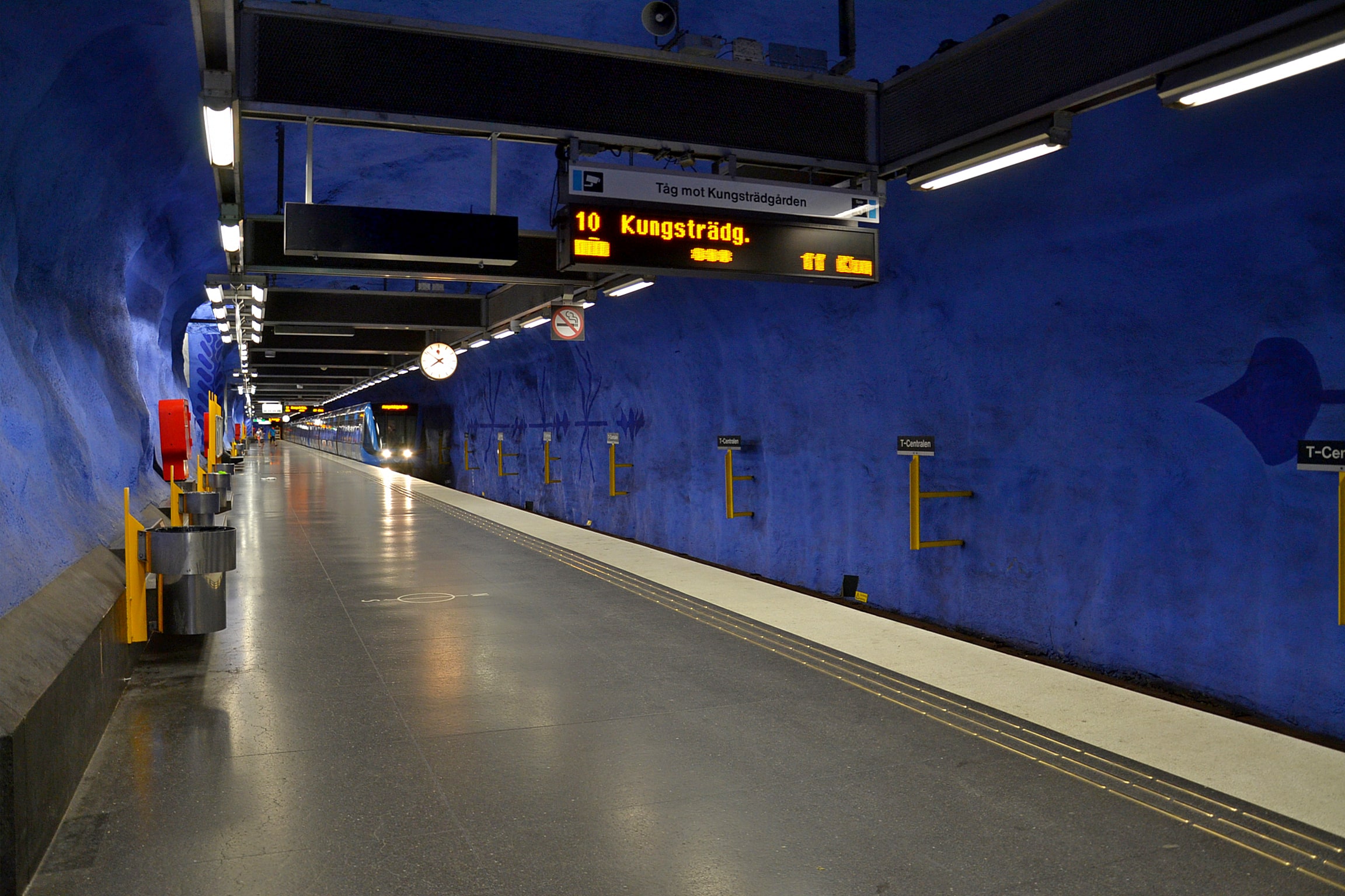 A blue-walled platform at T-Centralen metro station in Stockholm with a train approaching in the distance.