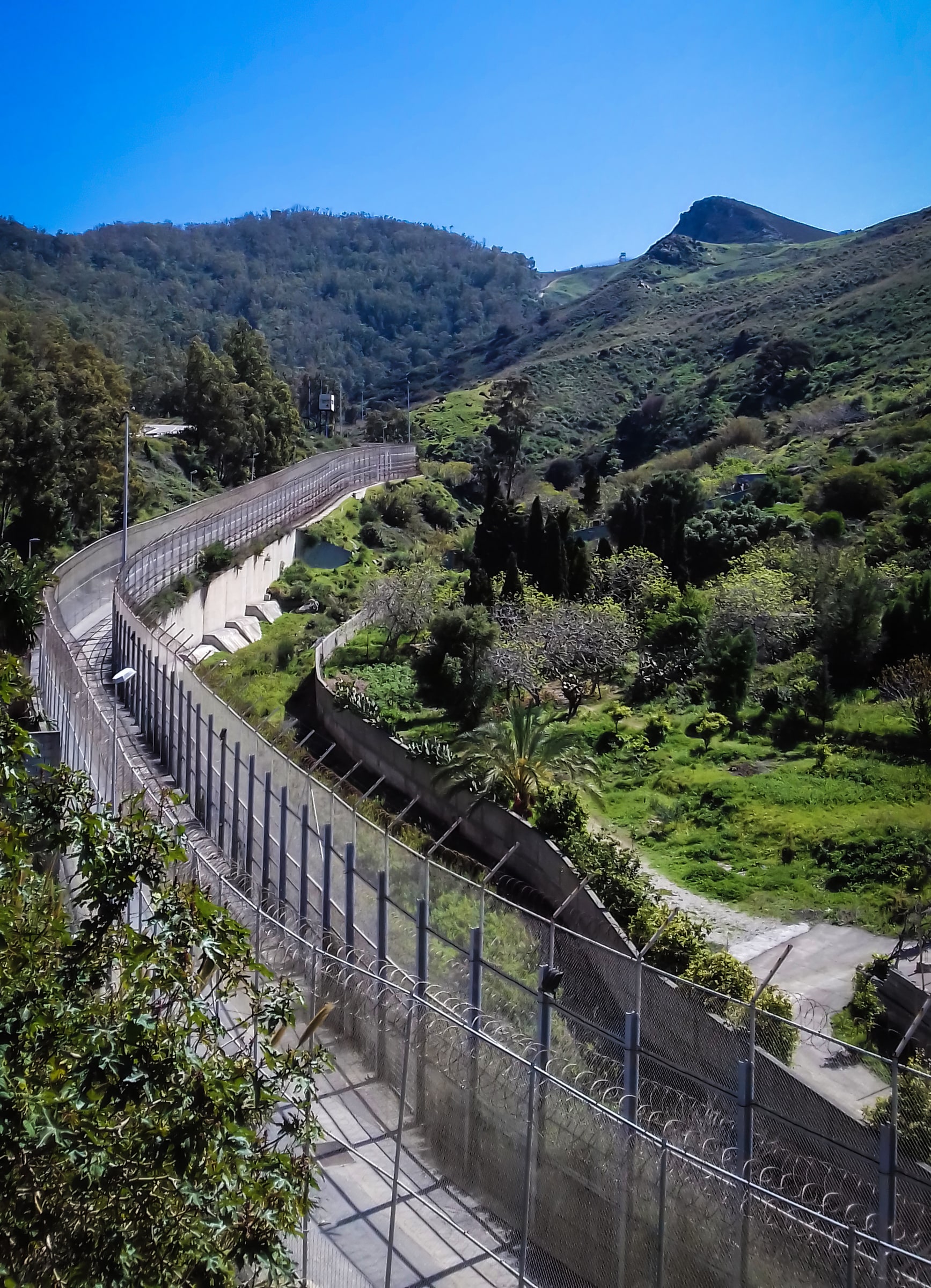The Ceuta-Morocco border fence winds through green hills on the Ceuta side.