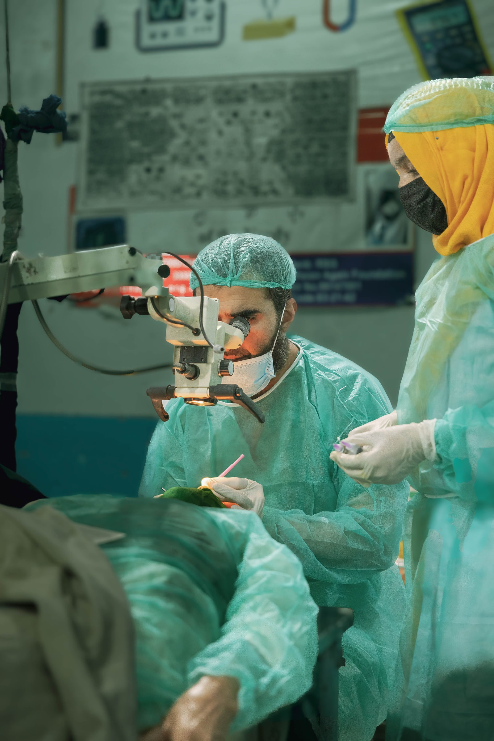 A masked surgeon uses an operating microscope while an assistant stands beside a draped eye-surgery patient.