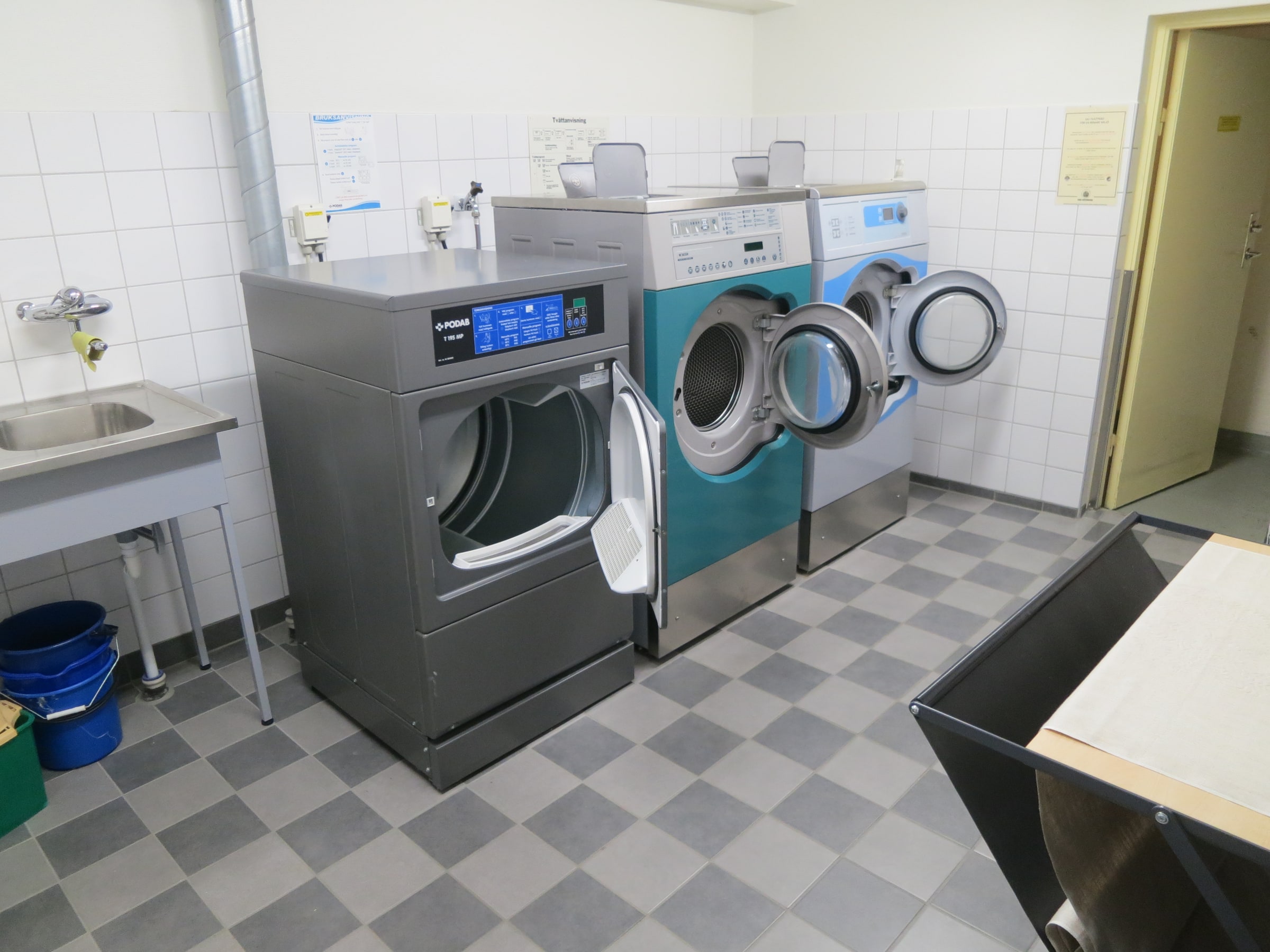 Two washing machines and a tumble dryer stand in a condominium laundry room in Gothenburg.