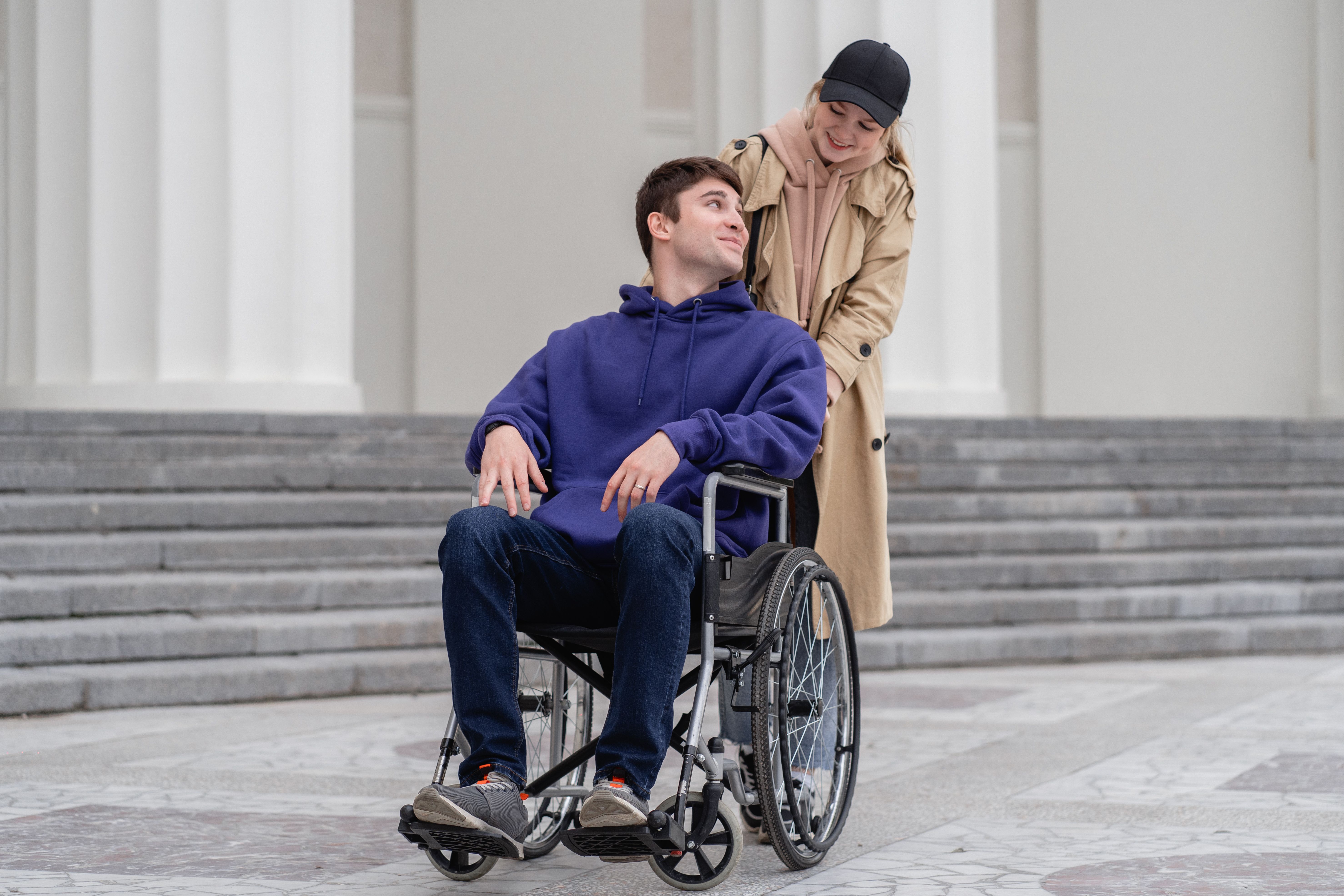 A woman pushes a smiling young man in a wheelchair outside a building.