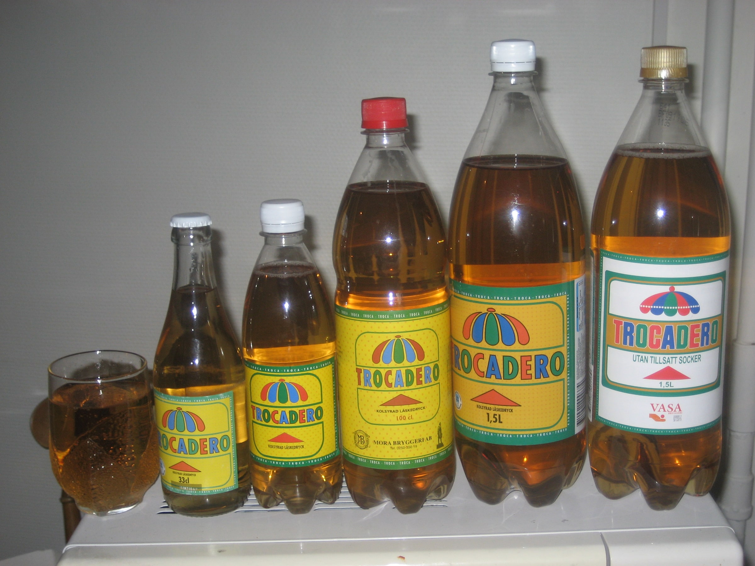 Five older Trocadero bottles in different sizes beside a glass of the soft drink.