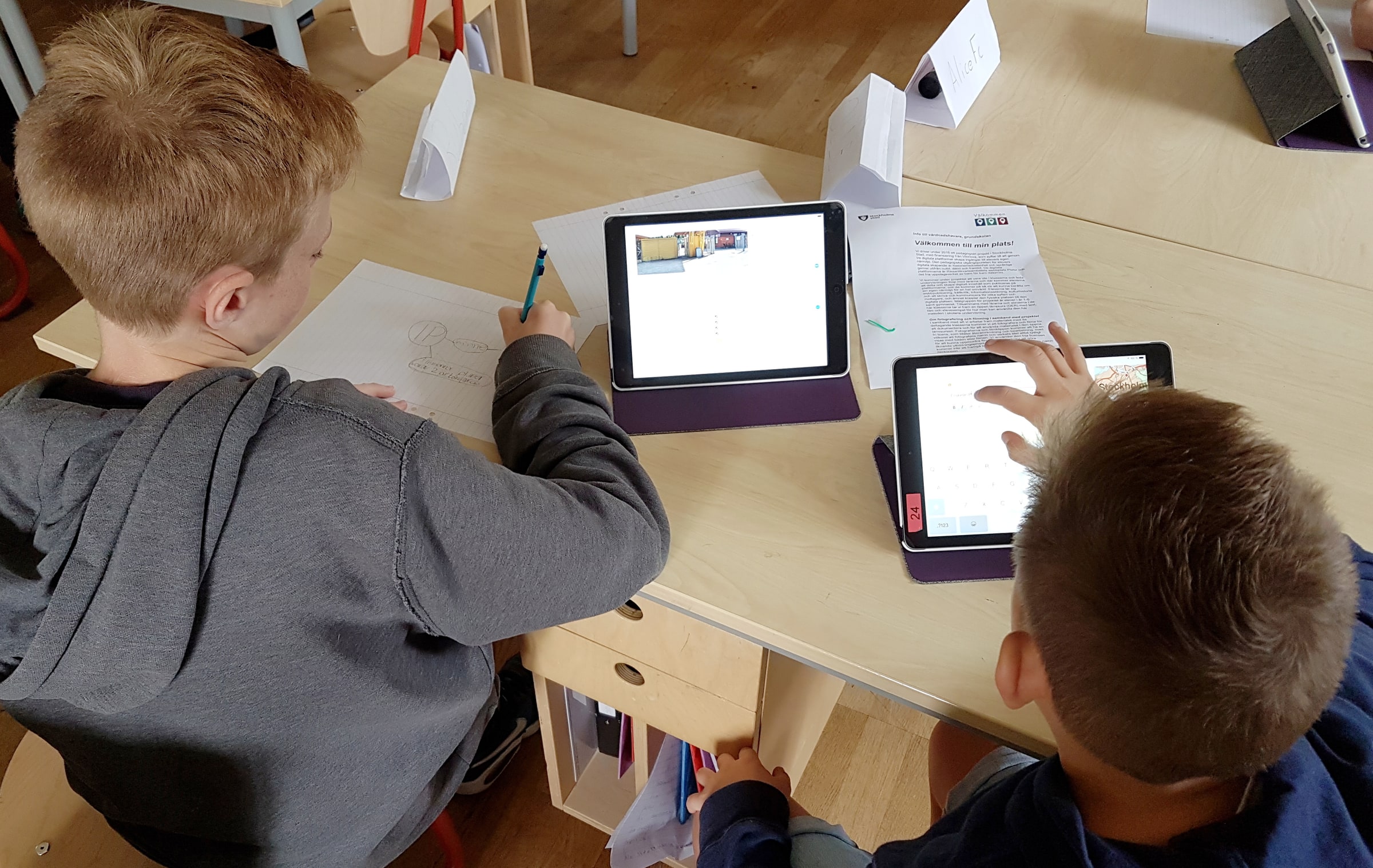 Pupils viewed from behind use paper, pencils and two tablets on a classroom table.