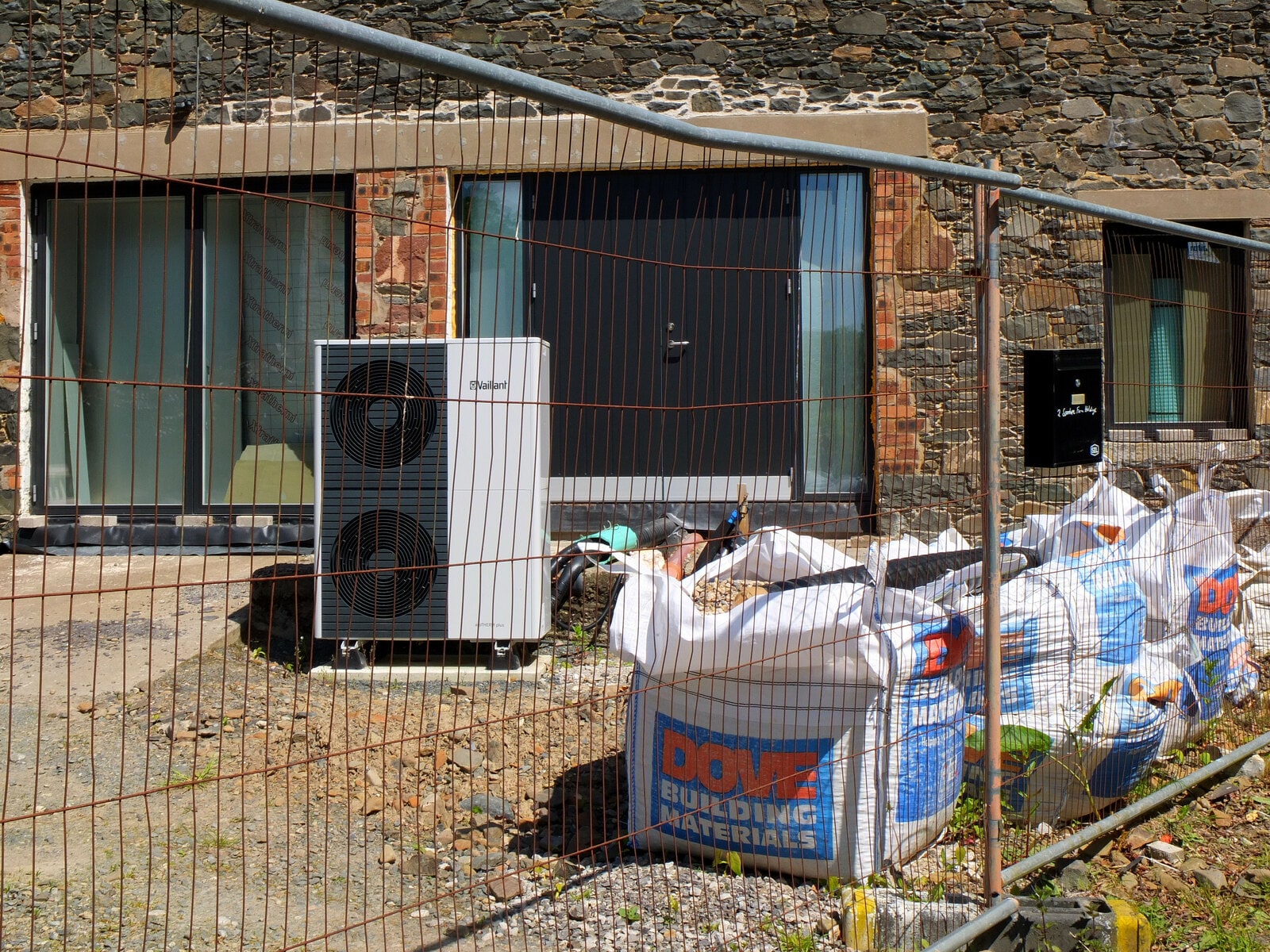 A heat-pump unit and building materials behind fencing outside a terraced home under renovation.