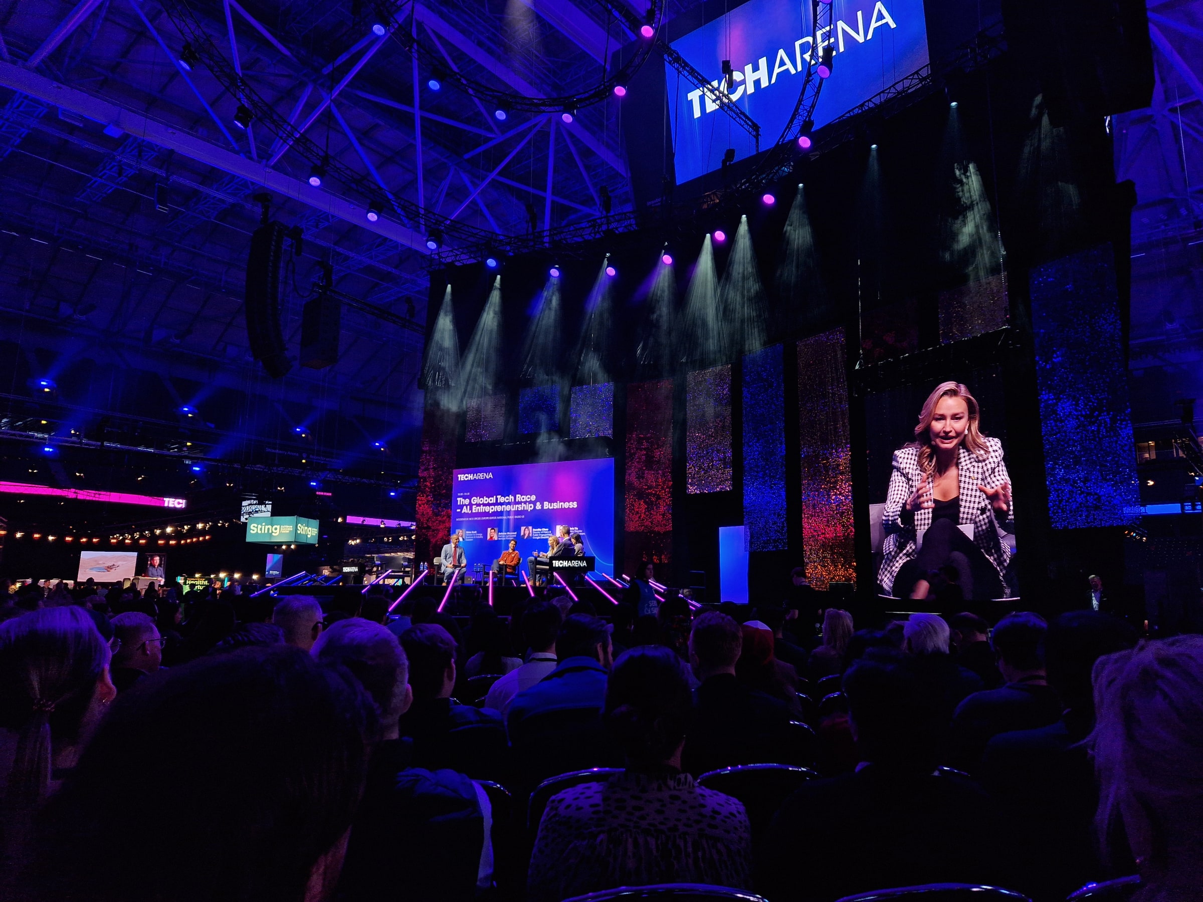 Ebba Busch speaking on a screen beside a conference stage and seated audience at Techarena in Stockholm.