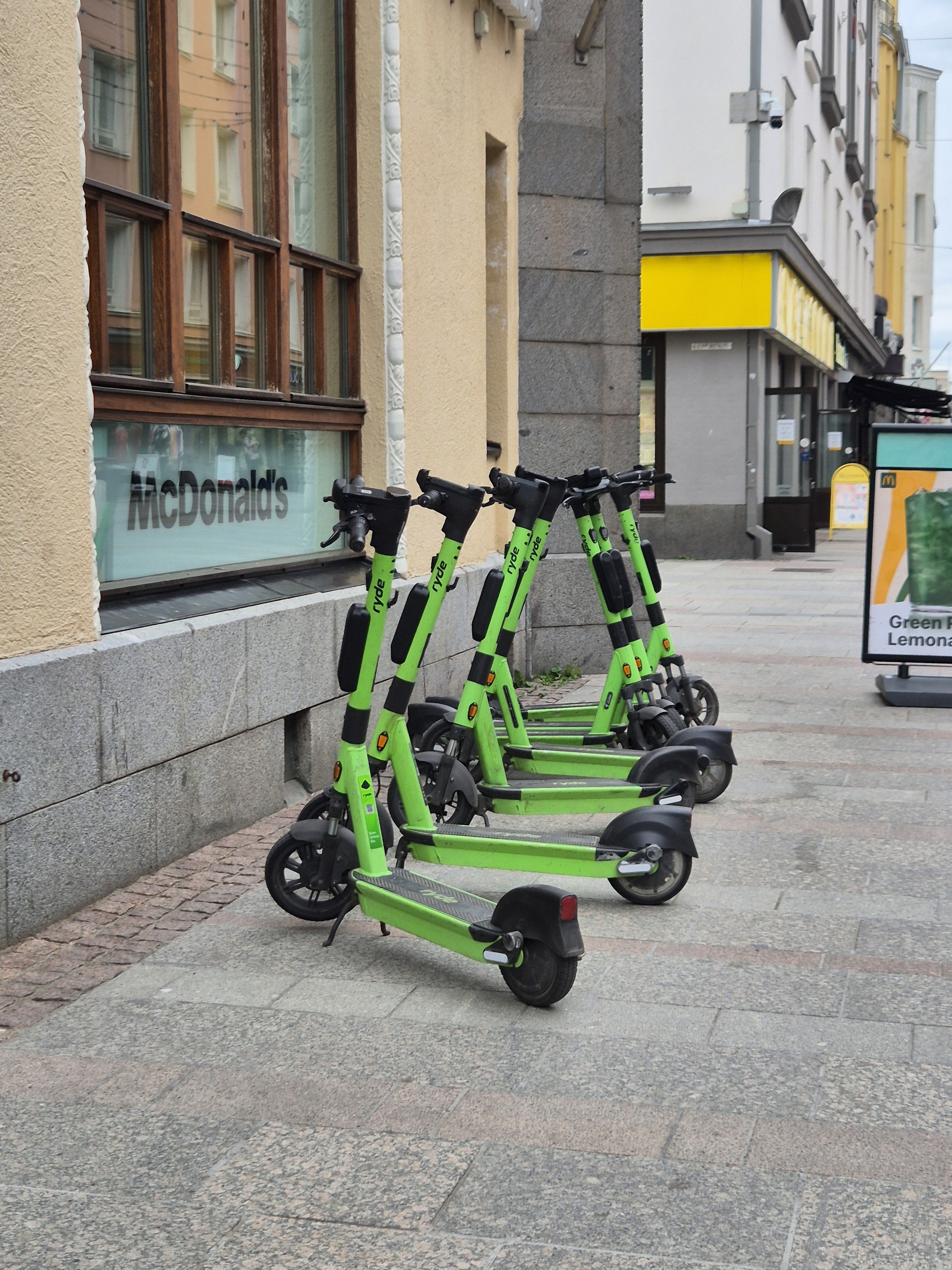 A row of green Ryde electric scooters is parked along a city pavement.