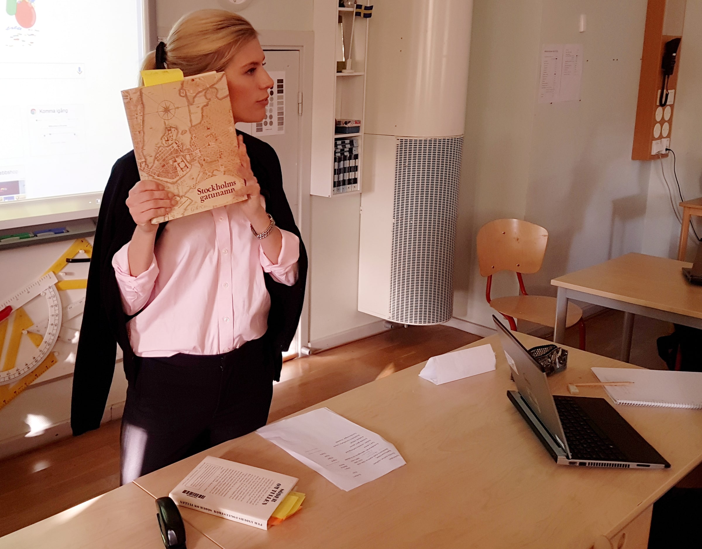 A school librarian holds up a book beside written lesson material and a laptop.
