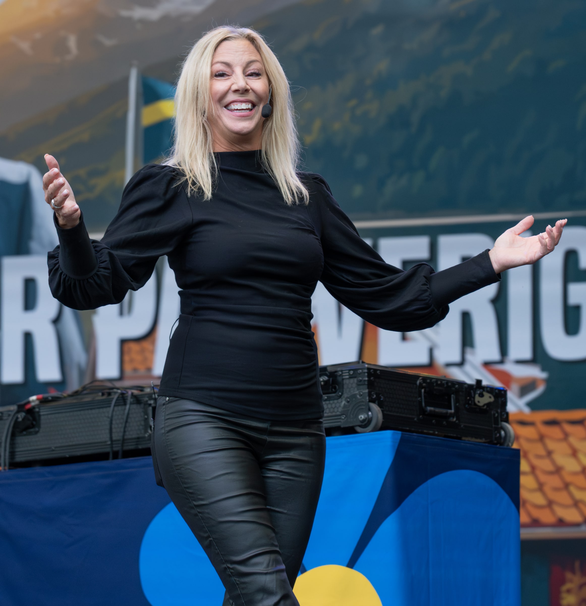 Linda Lindberg speaking with a headset microphone and open hands on a stage in Stockholm.