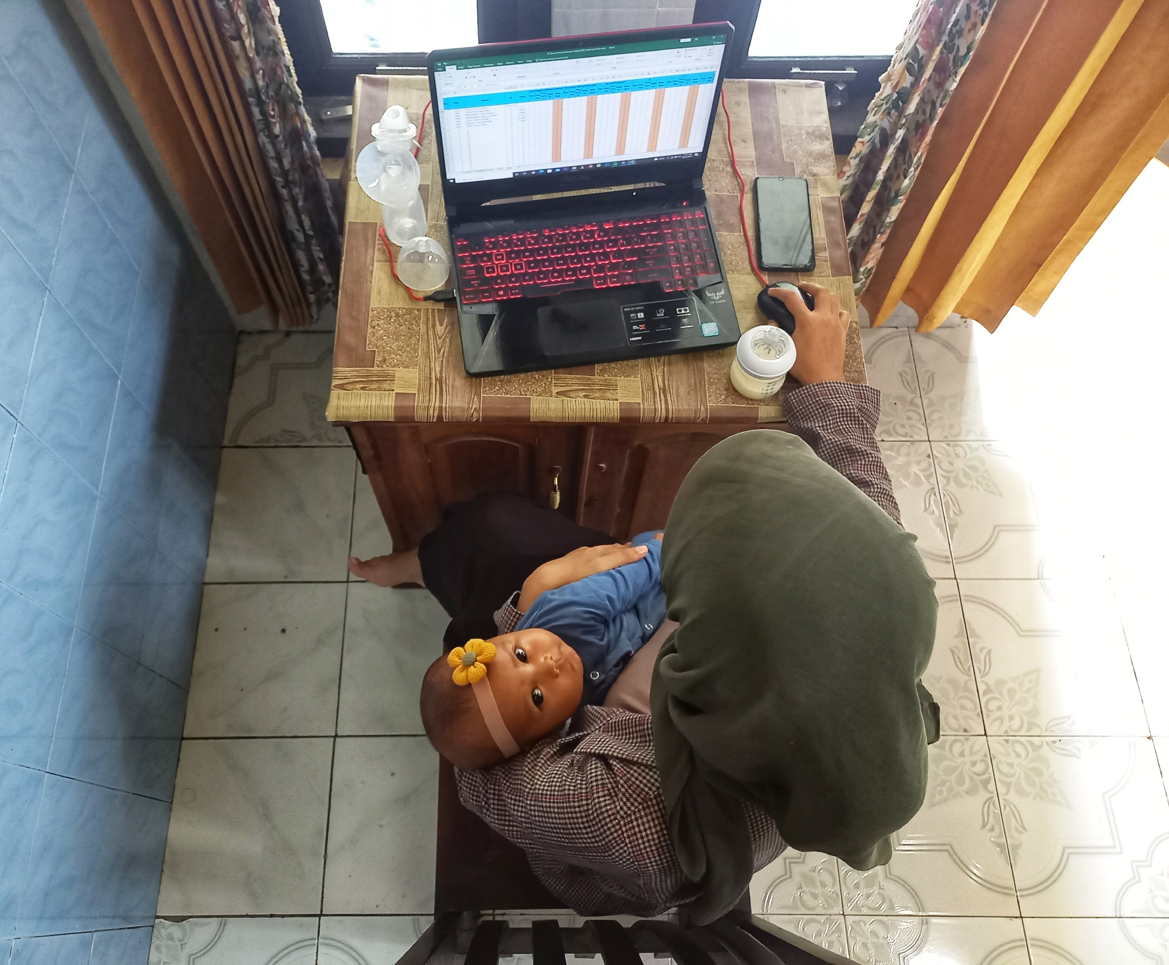A woman works on a laptop while an infant rests across her lap beside feeding equipment.