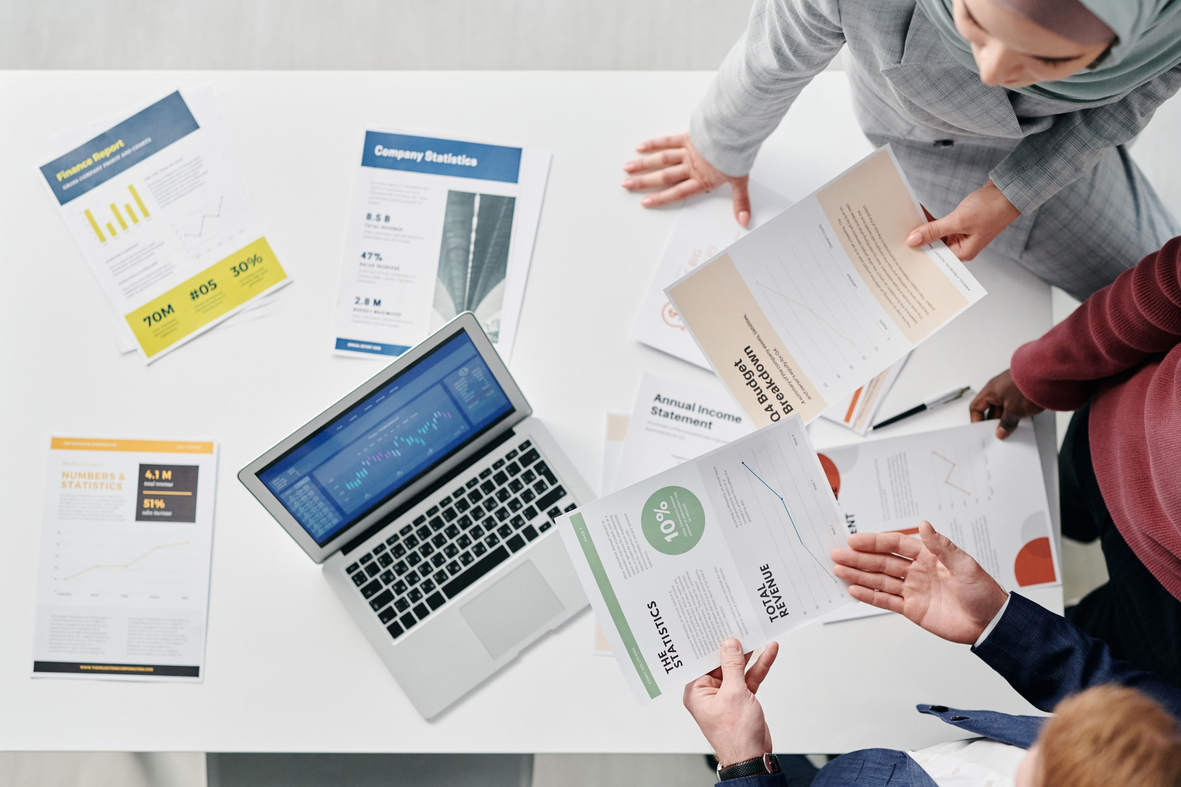 People compare printed reports and charts around a laptop at a meeting table.