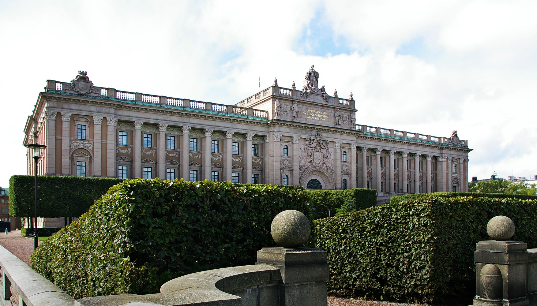 The eastern facade of the Swedish Parliament building in Stockholm behind trimmed hedges.