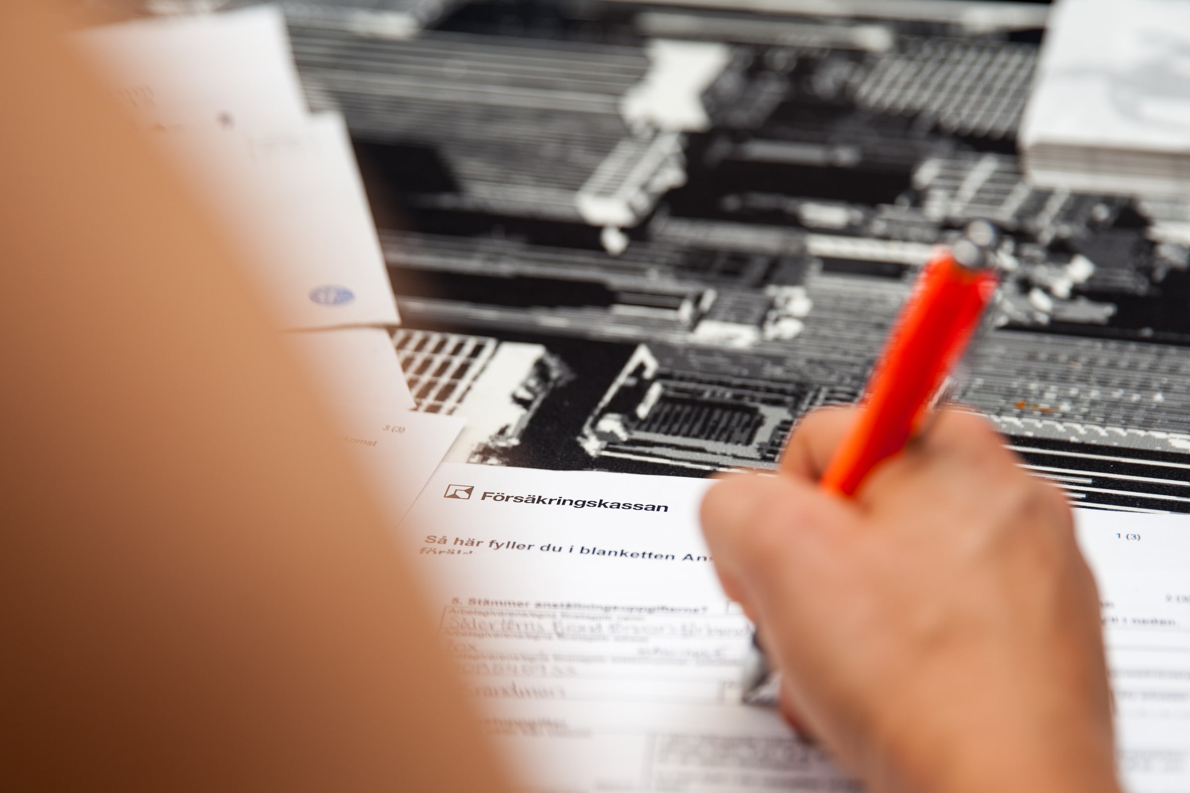 A hand holds a red pen over Försäkringskassan paperwork beside a laptop.