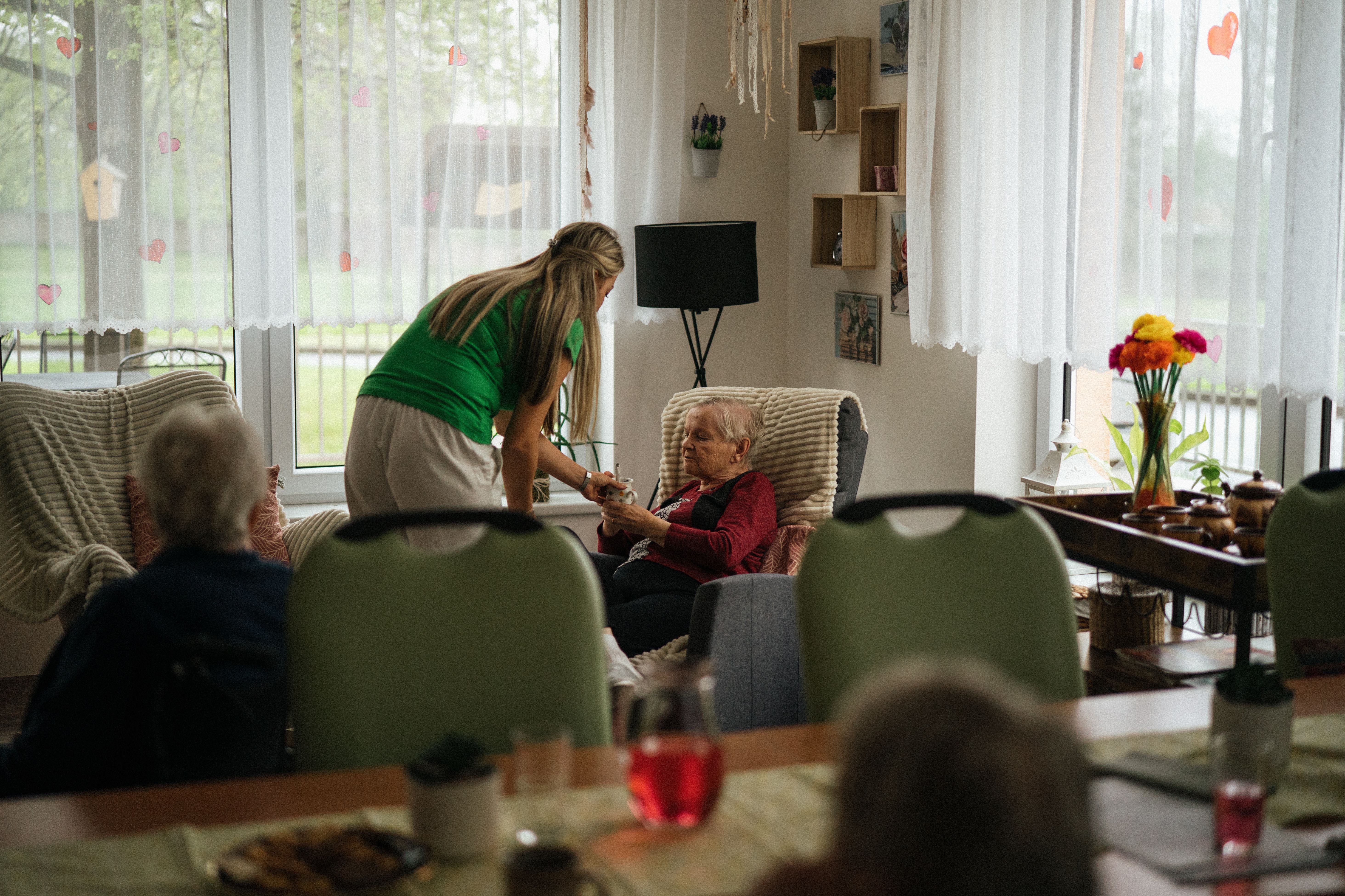 A caregiver hands a cup of tea to an older woman seated in a care home.