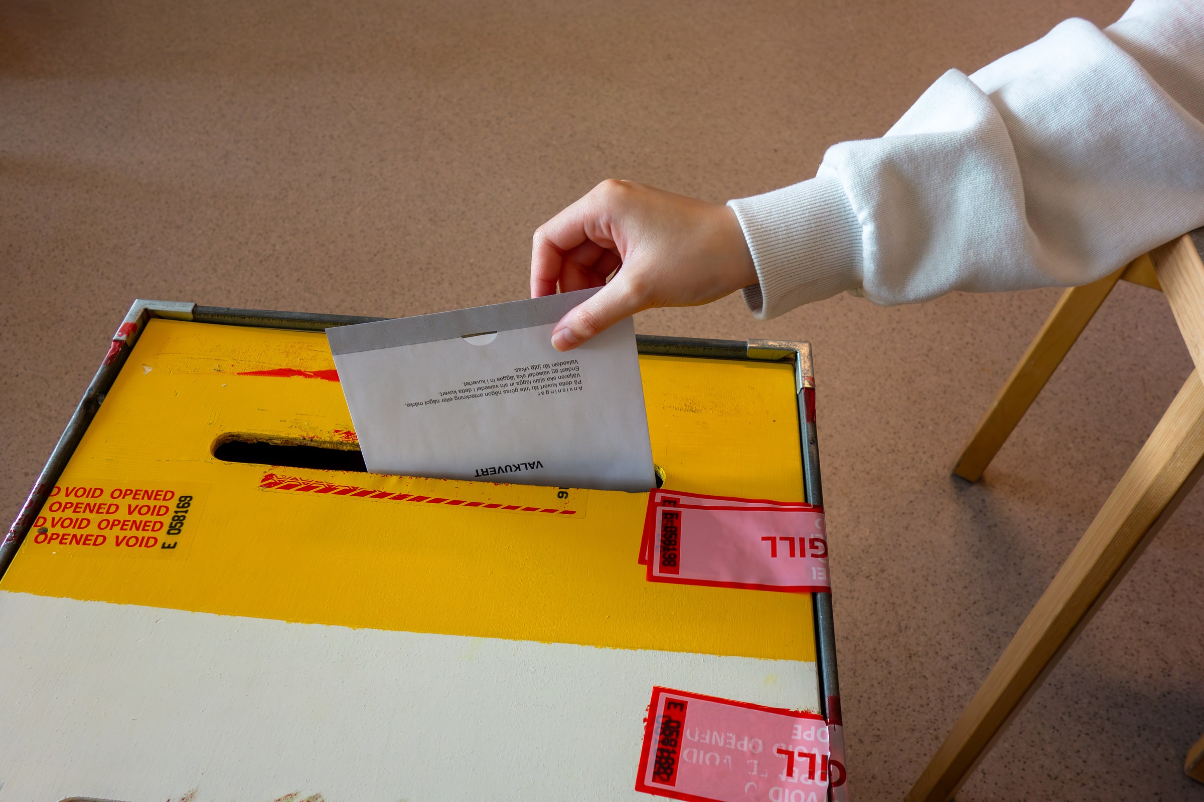 A hand places a ballot envelope into a sealed ballot box at a Swedish polling station.