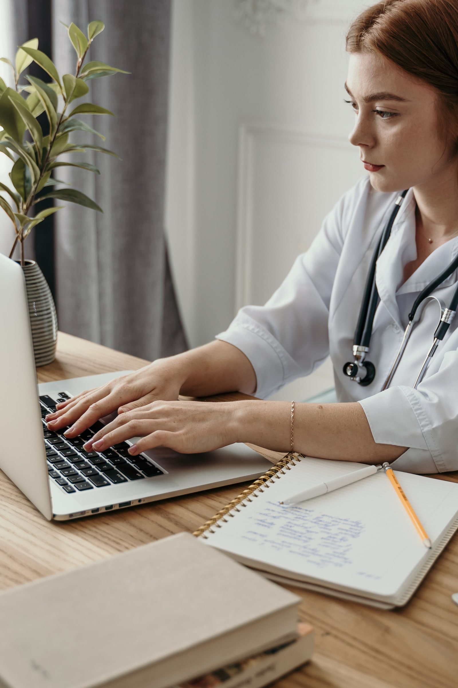 A clinician in a white coat types on a laptop beside a stethoscope and handwritten notes.