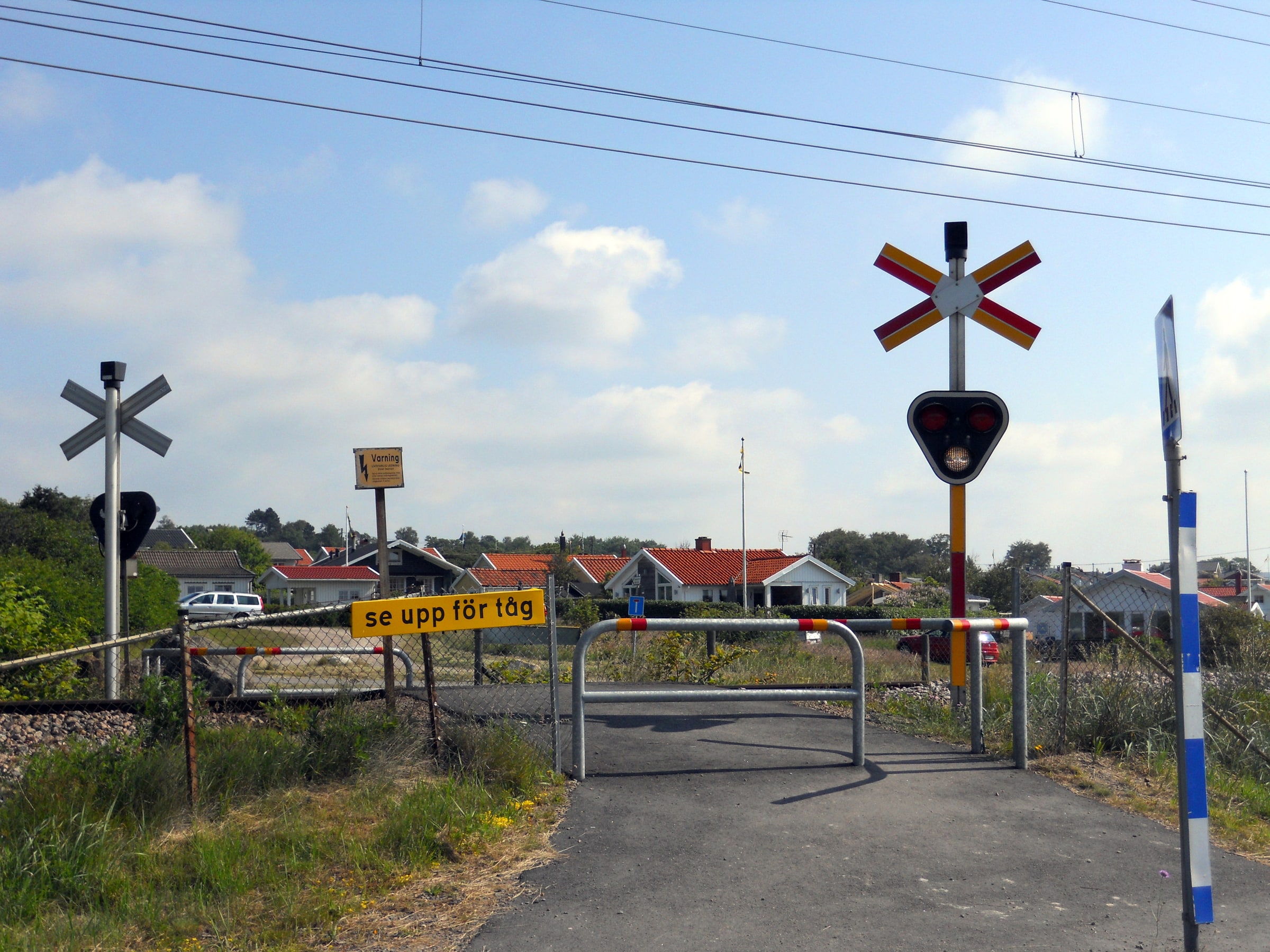 A marked pedestrian crossing over railway tracks at Apelviken in Varberg, with barriers, warning lights and a sign telling people to watch for trains.