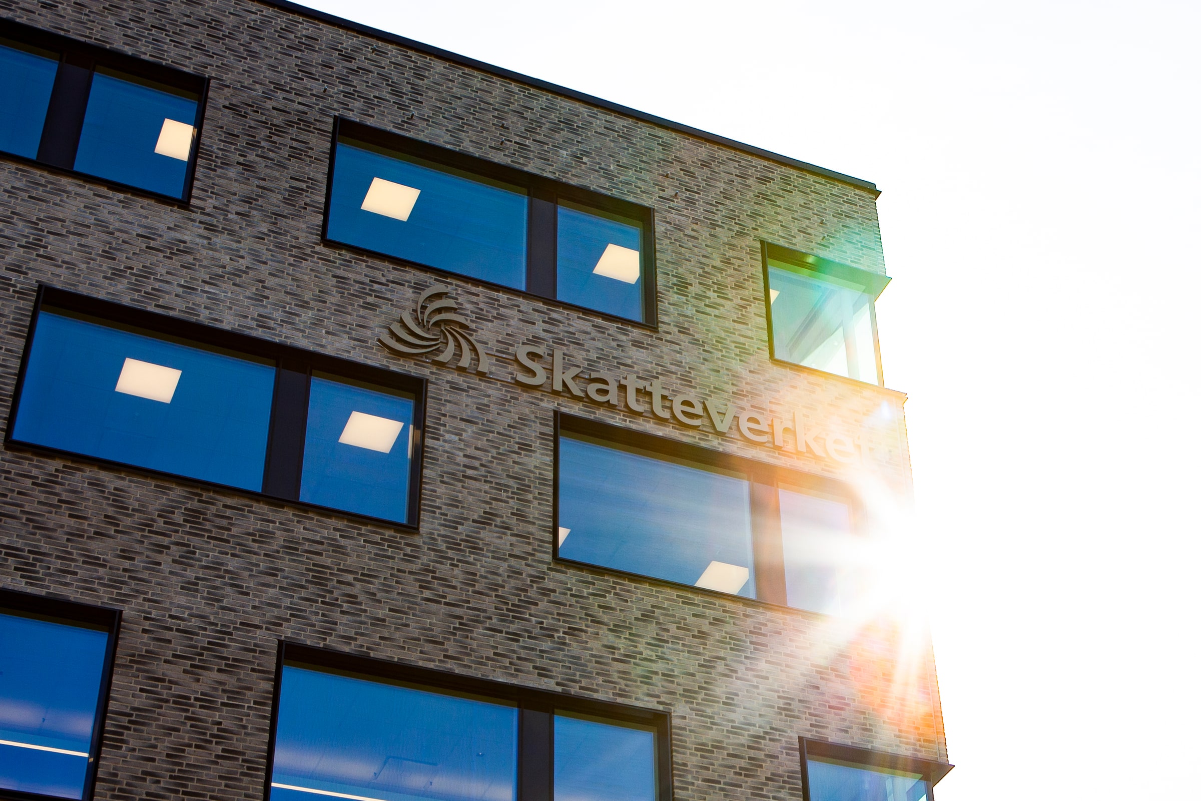 The Skatteverket name and symbol mounted on the brick facade of the Kronan office in Sundbyberg, lit by low sunlight.