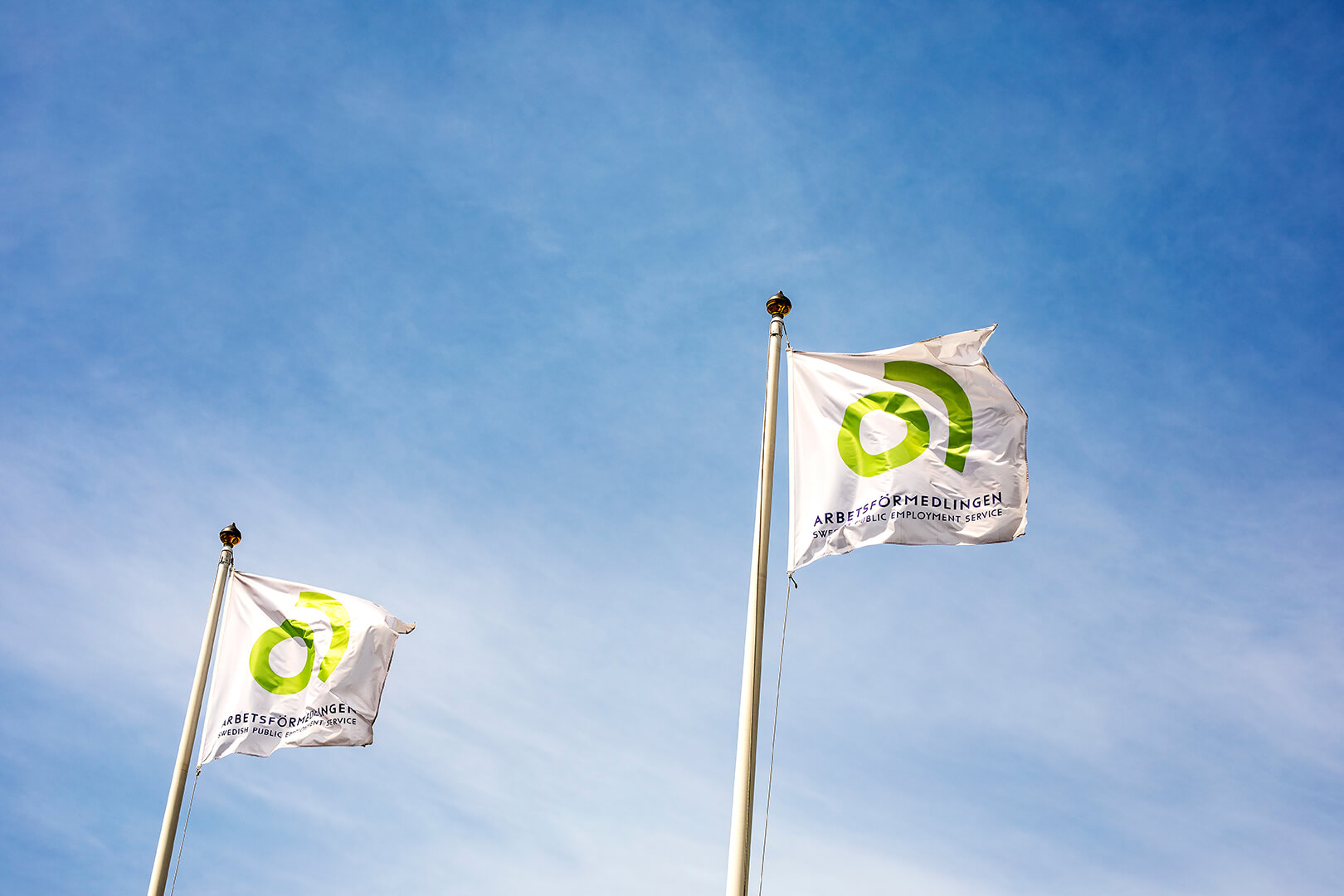 Two white Arbetsförmedlingen flags flying on tall poles against a blue sky.
