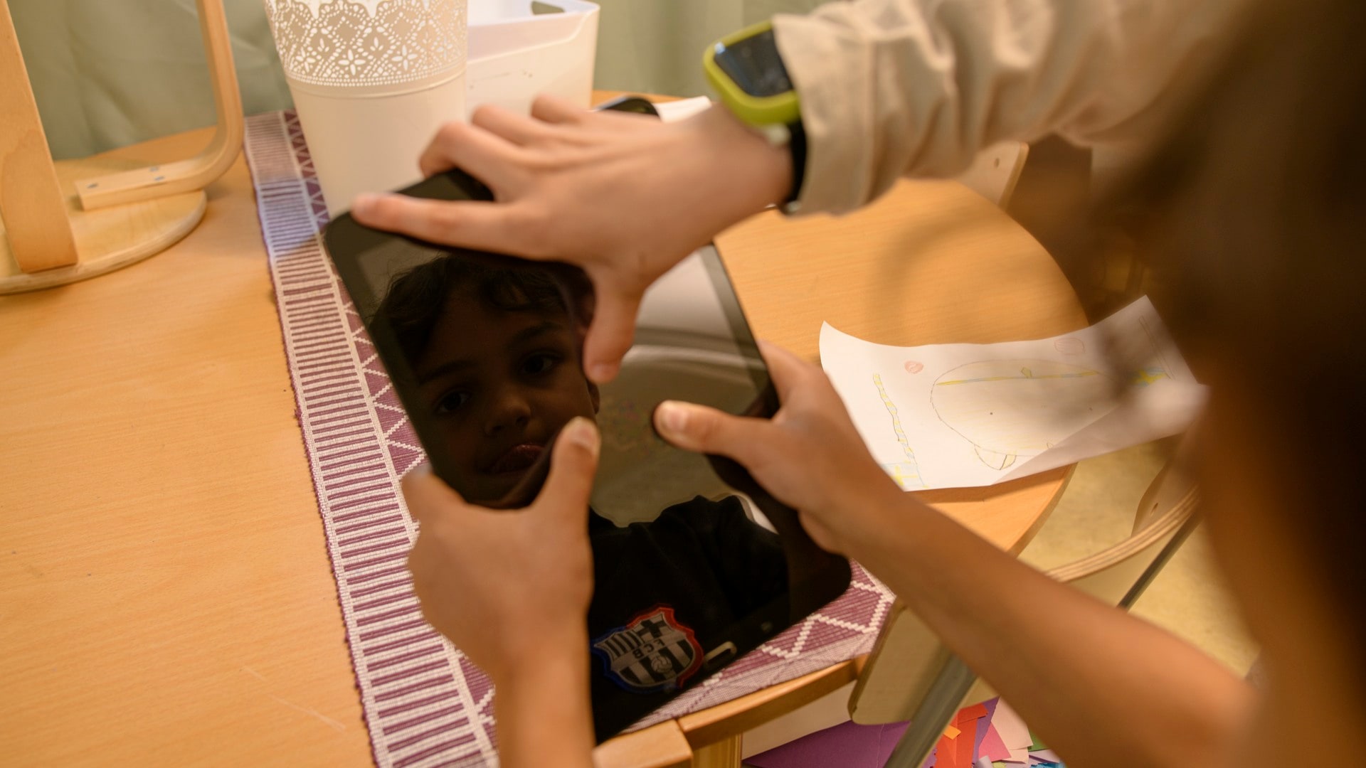 Children hold a black tablet above a table, with a face reflected in its screen.