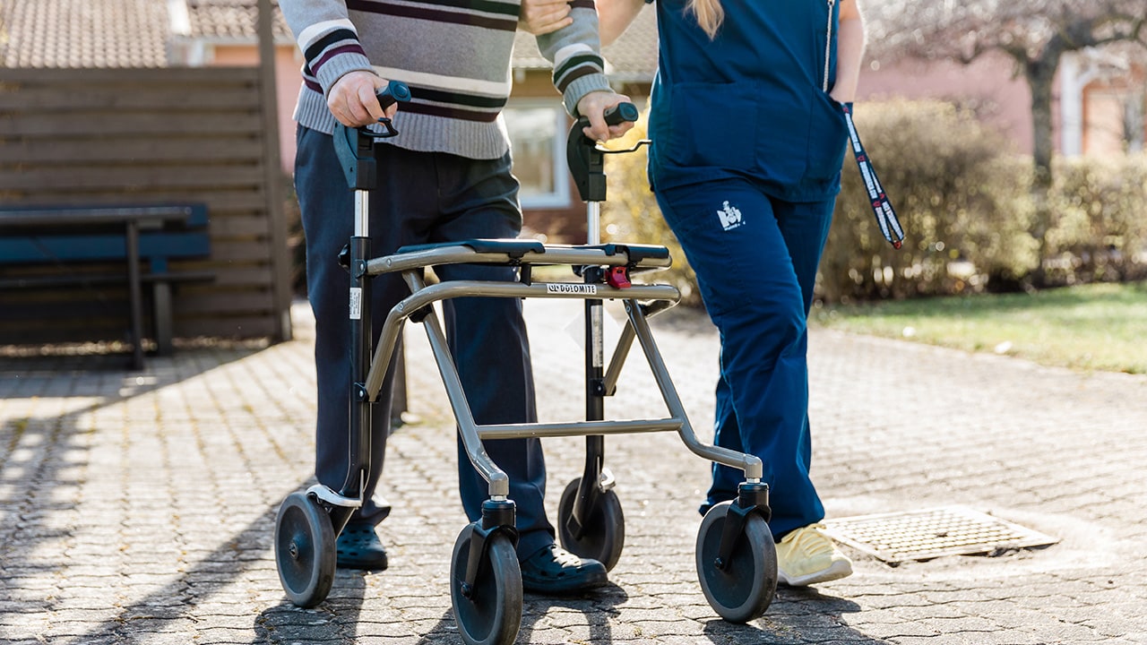 A care worker walks beside an older person using a wheeled walker outdoors in Malmö.