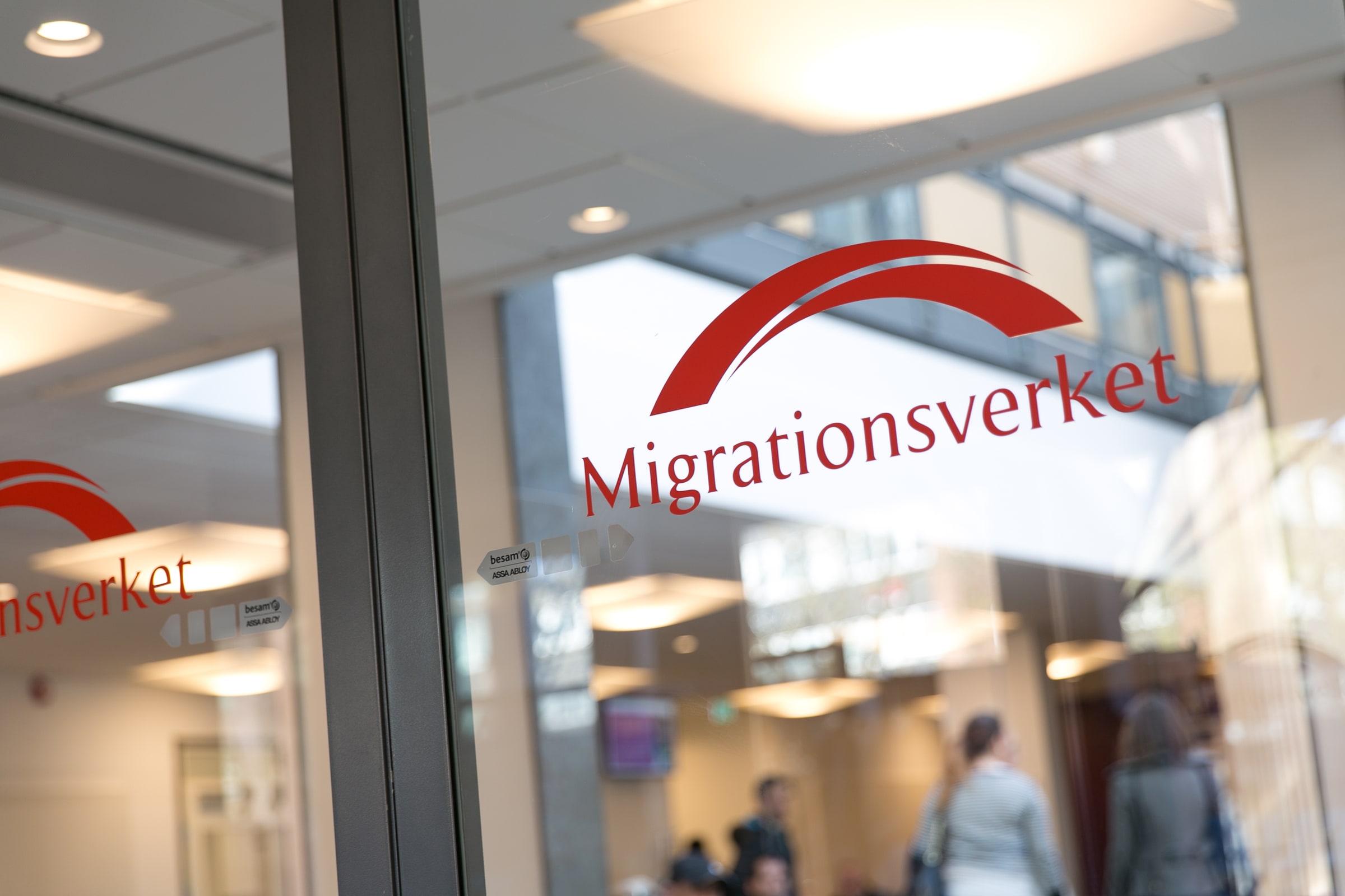 Migrationsverket's red logo on a glass wall inside a Swedish Migration Agency office.