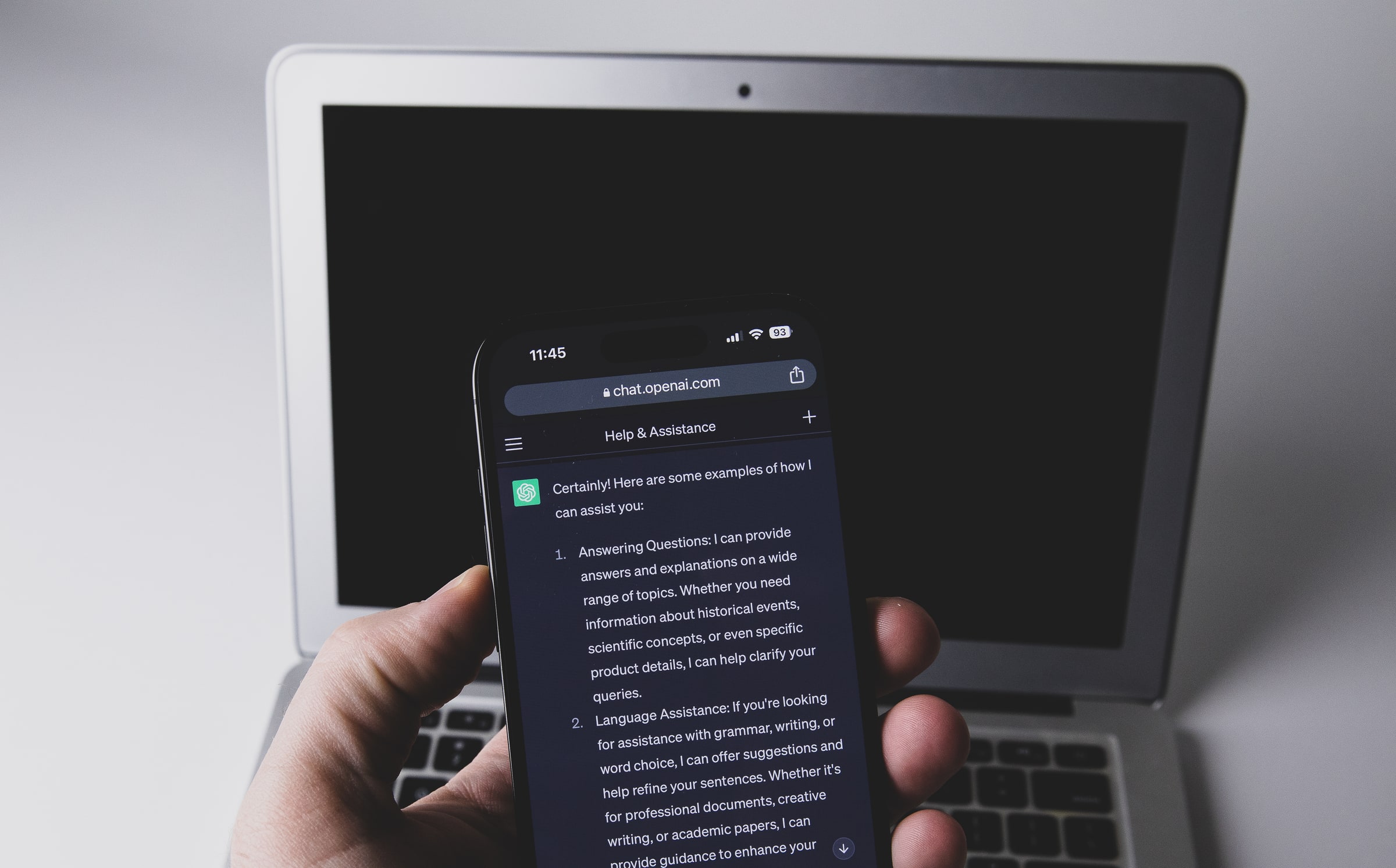 A hand holds a phone displaying a ChatGPT response in front of a laptop, May 2023.