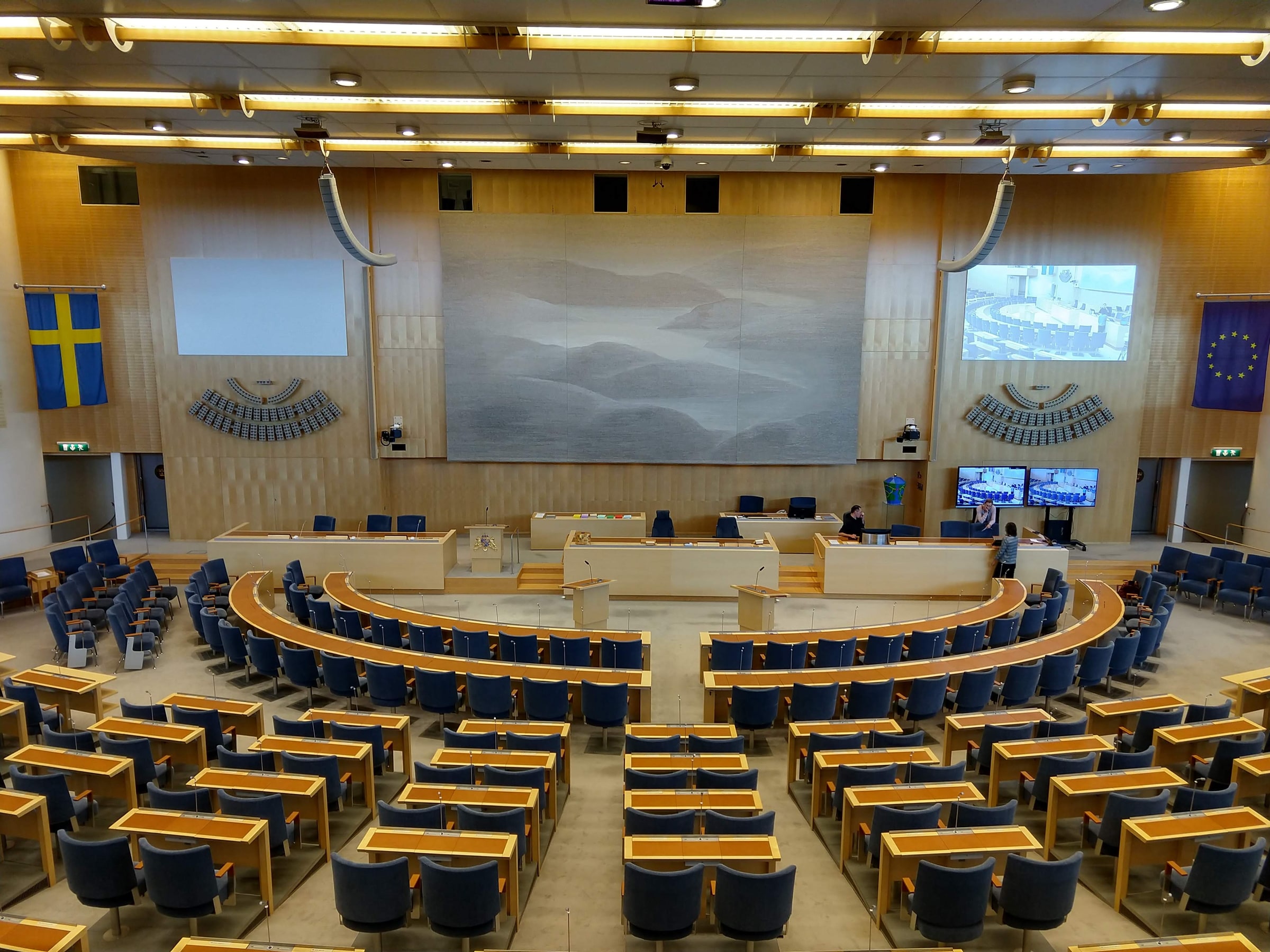 The Swedish Parliament chamber with blue seats, central lecterns and Swedish and EU flags.