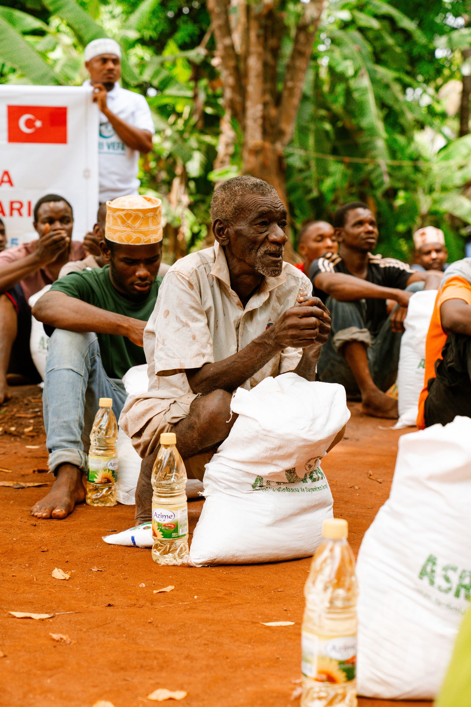 People sit beside food sacks and cooking oil during an outdoor aid distribution.